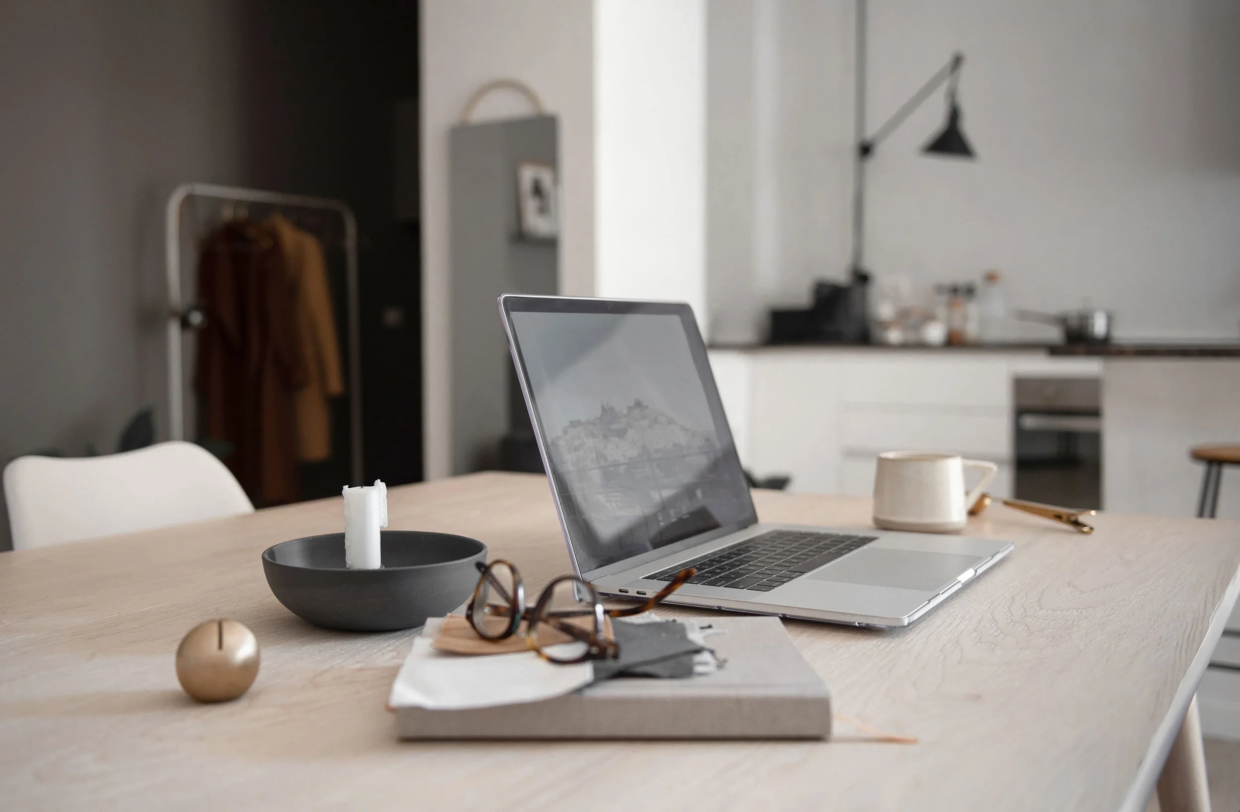 A wooden dining table with a silver laptop, a black bowl with a white candle, a pair of glasses, and a beige mug. In the background, there is a kitchen with white cabinets, a black floor lamp, and a coat rack with brown coats.