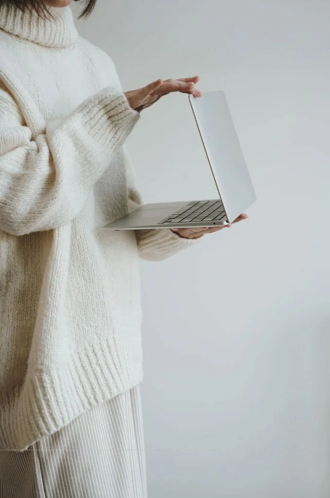 A person wearing a cream-colored sweater holding a silver laptop with a blank white screen against a plain white background.