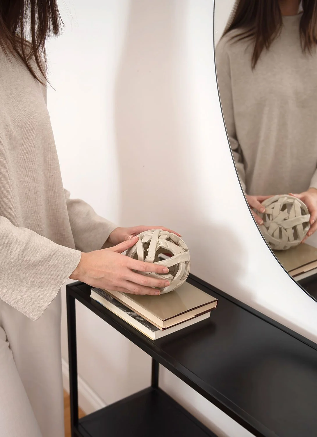 A person in beige places a decorative ball on books beside a round mirror on a black console table.