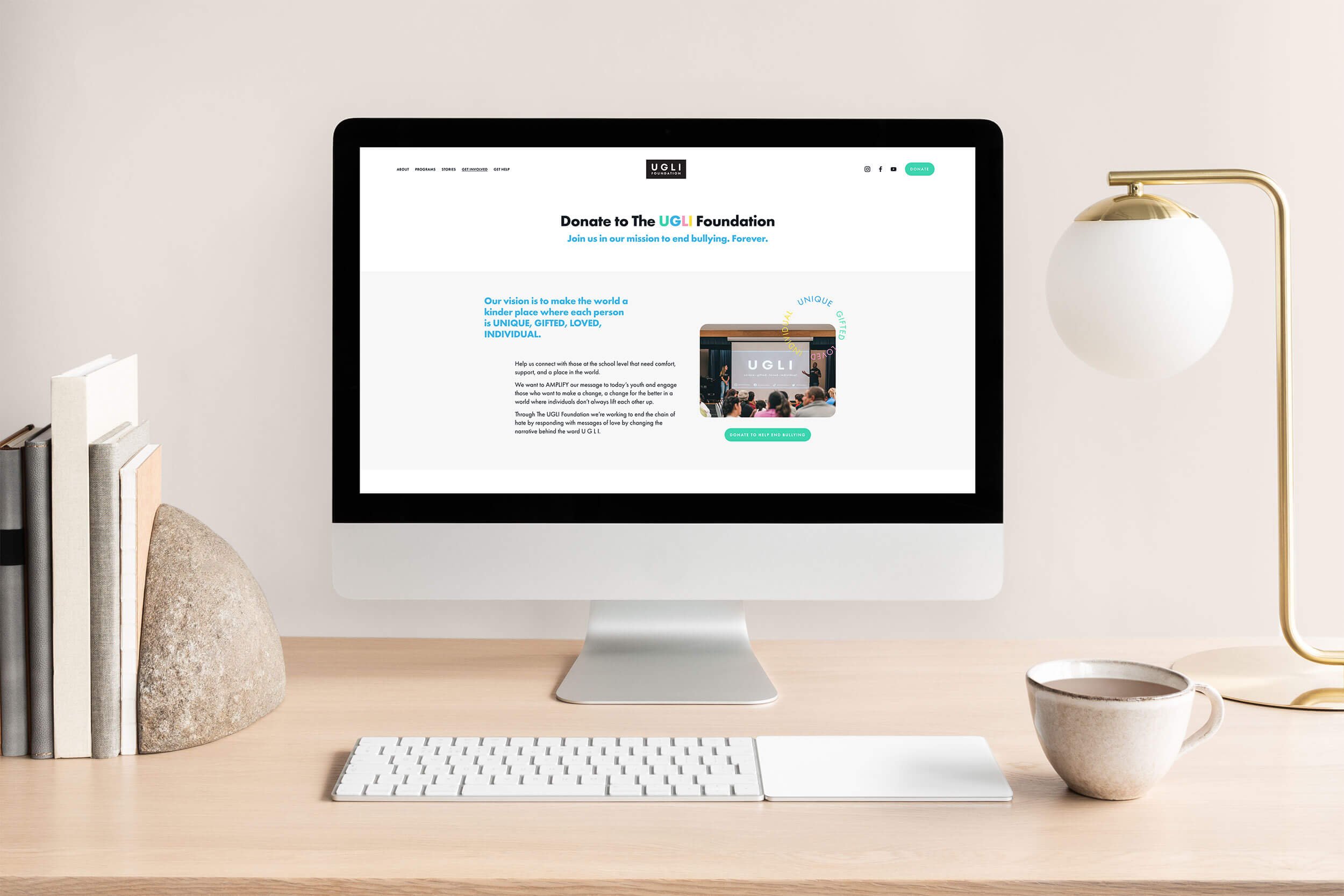 Computer monitor on a desk showing the UGLI Foundation website, with books, a lamp, and a coffee mug nearby.