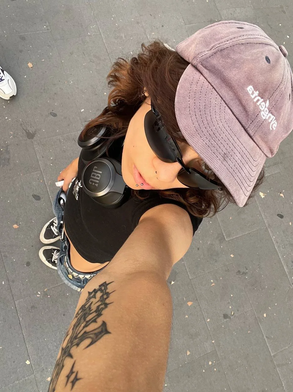A woman taking a selfie from above, wearing sunglasses, a pink baseball cap, black crop top, denim shorts, and headphones, with a tattoo on her arm.