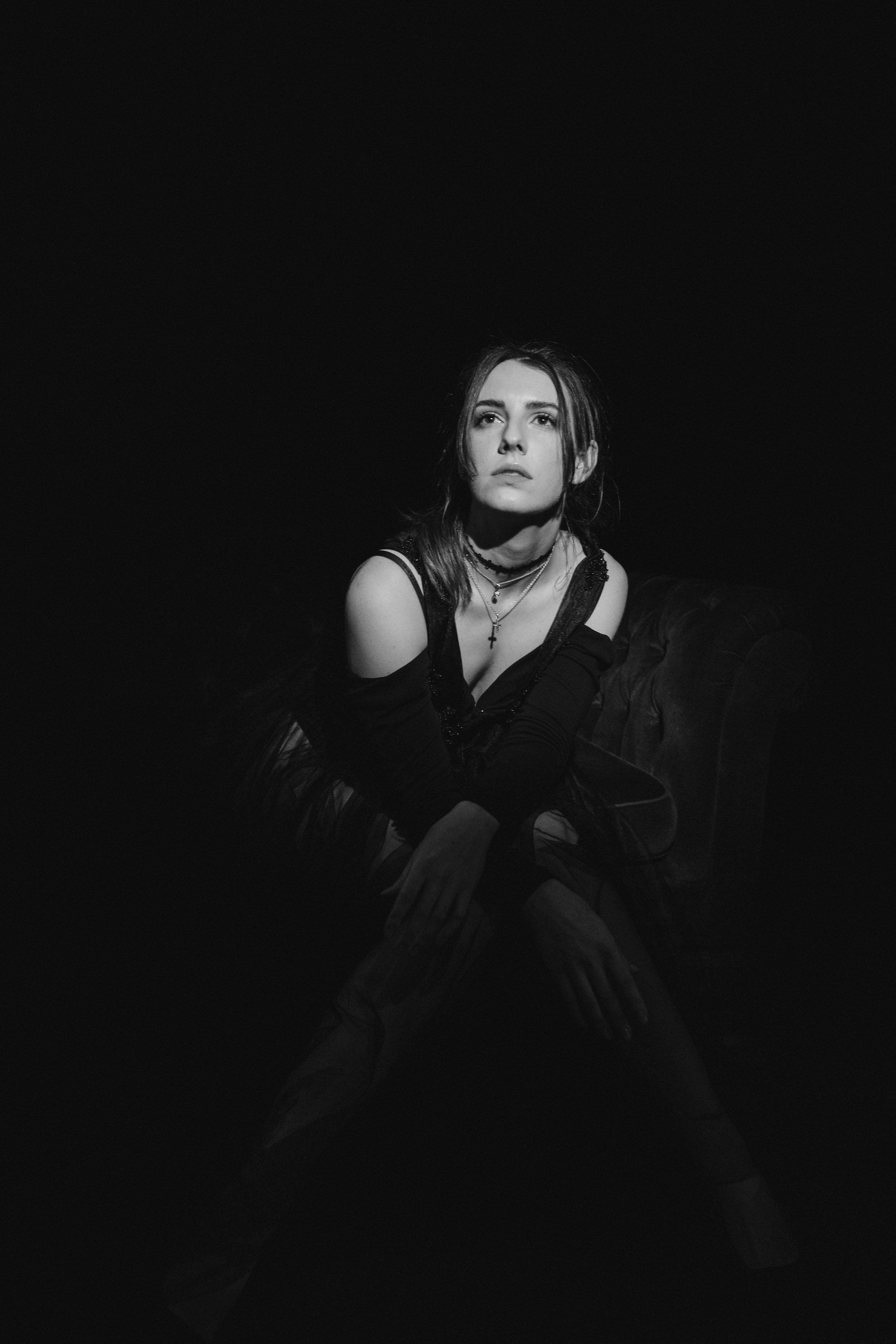 A woman dressed in dark clothing with layered necklaces sitting on a dark chair or sofa in a dimly lit setting with a black background.