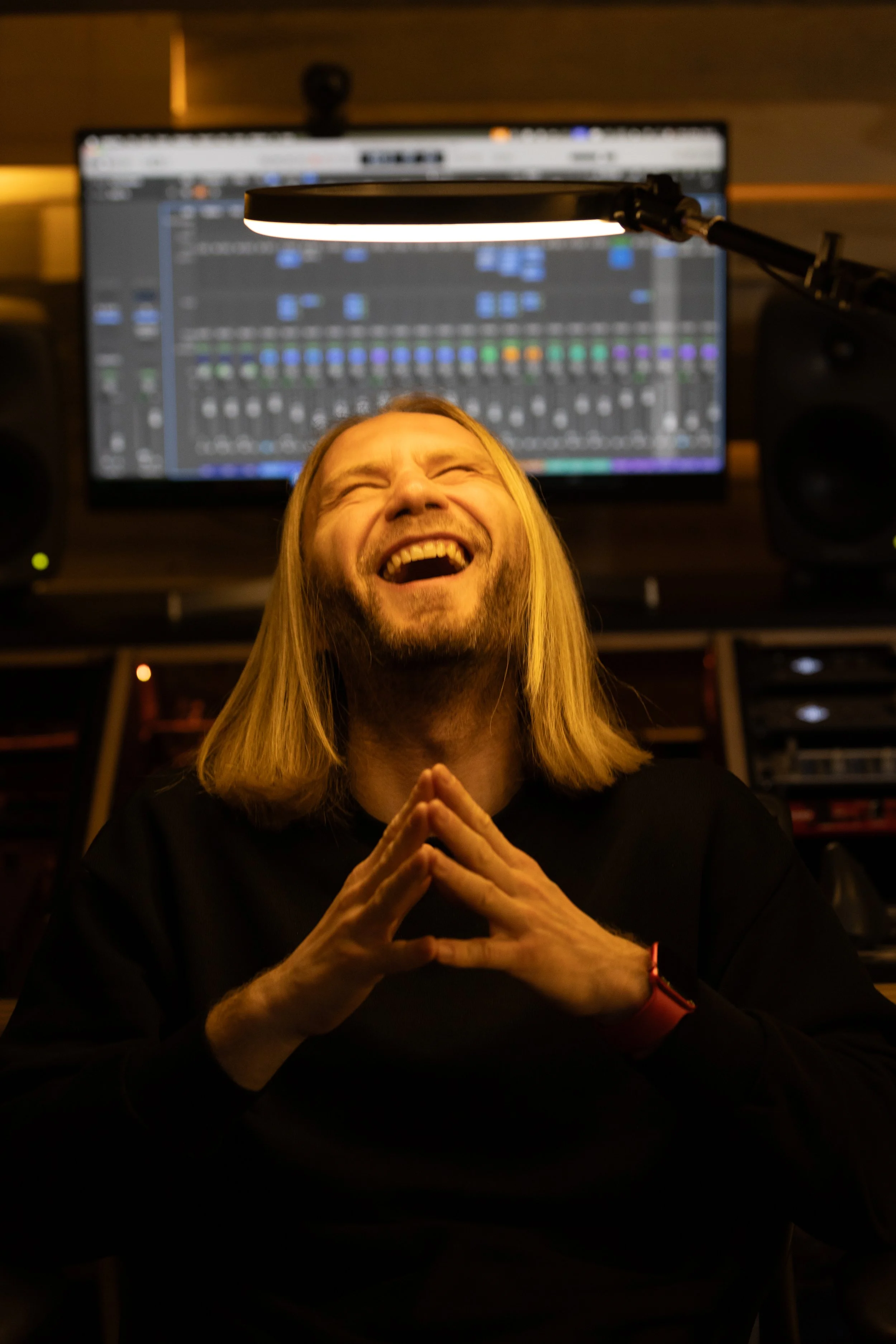 A man with long blonde hair laughing with closed eyes, hands clasped together, seated in a recording studio with a computer screen showing audio editing software in the background.