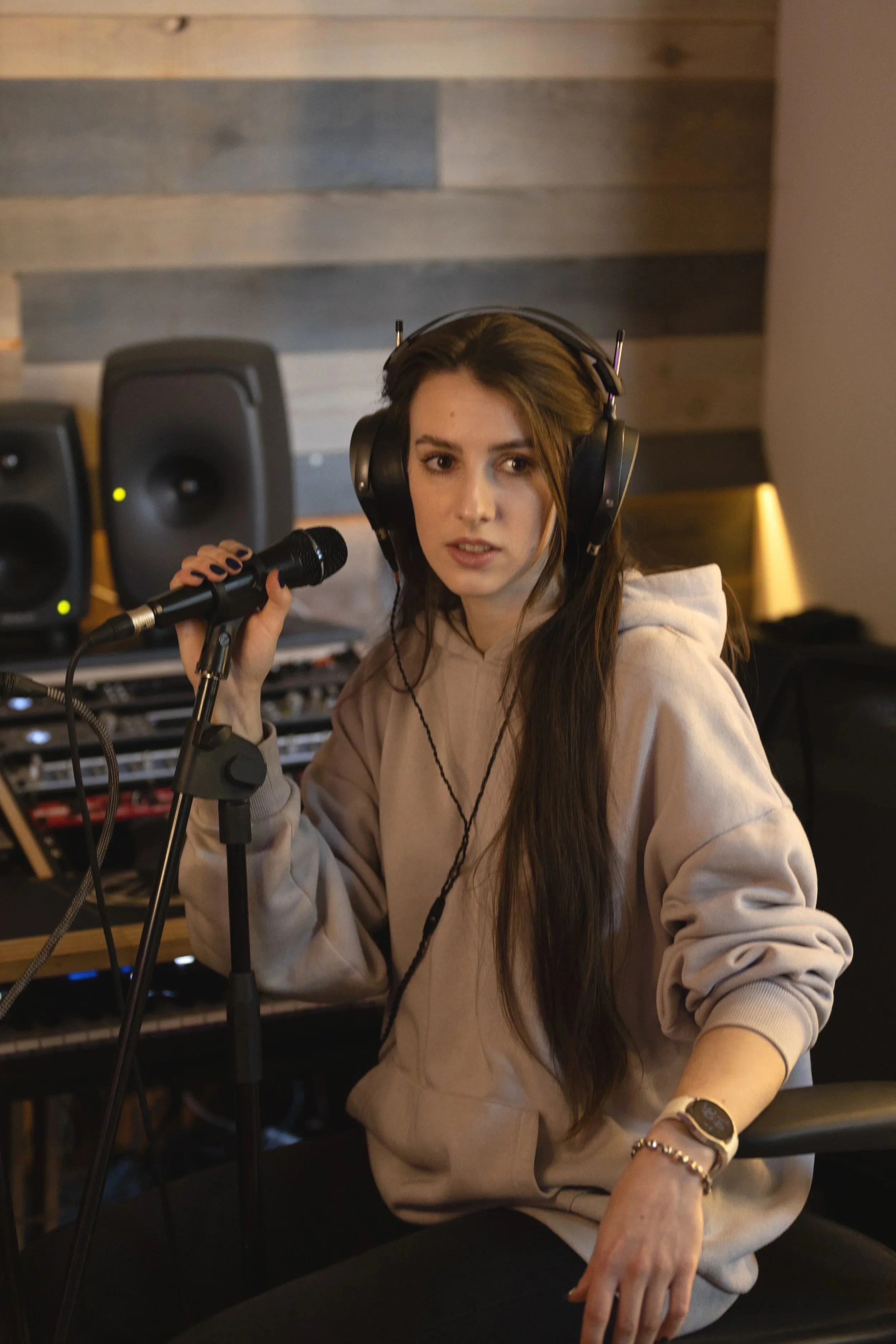 Woman in a beige hoodie sitting in a recording studio, holding a microphone, wearing large headphones, with audio equipment and speakers in the background.