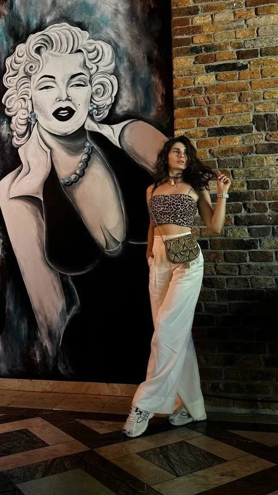 A woman with dark hair, wearing a leopard print crop top, white wide-leg pants, and sneakers, stands in front of a large black-and-white portrait of Marilyn Monroe on a brick wall.
