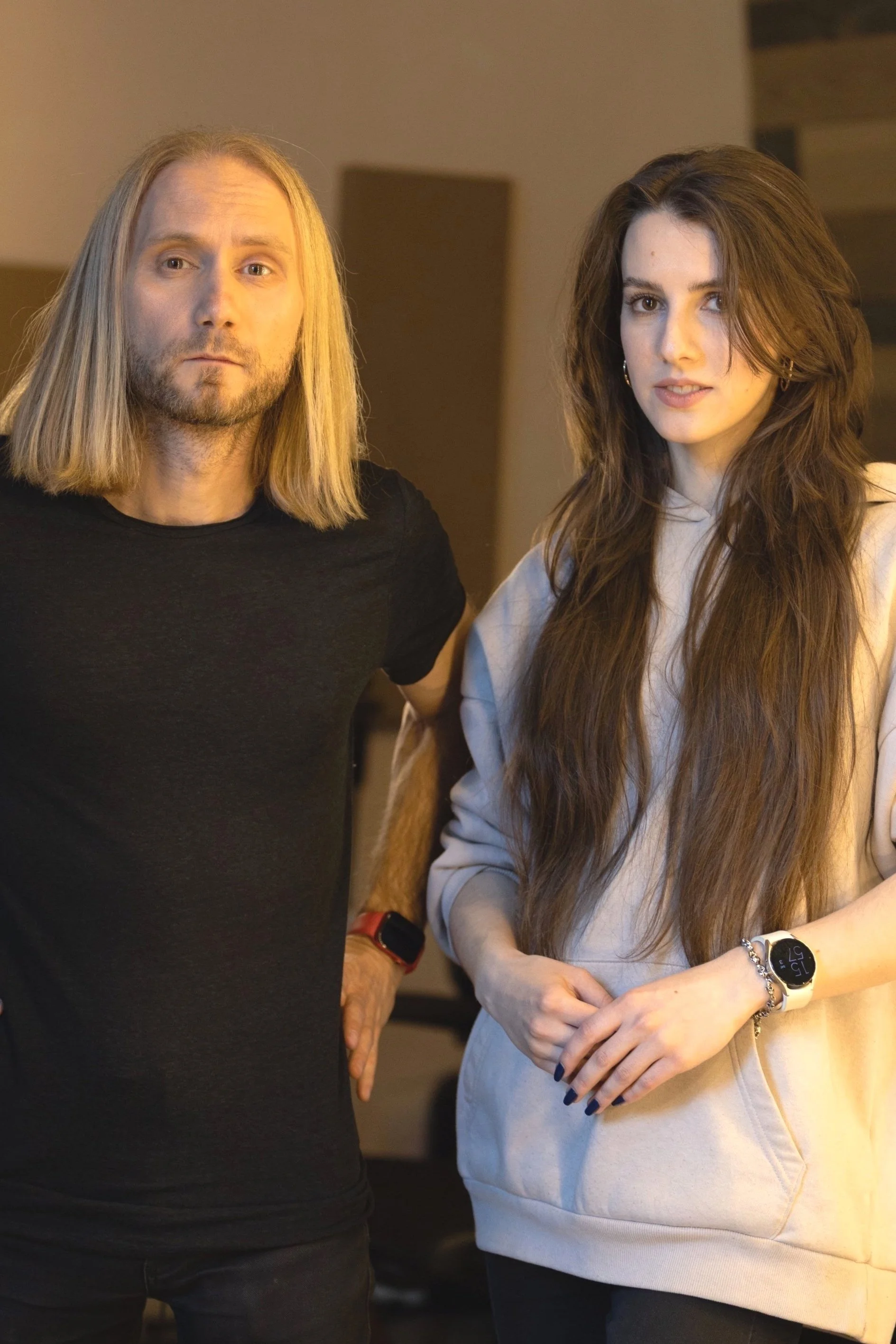 A man with long blond hair and a beard wearing a black t-shirt standing next to a young woman with long brown hair wearing a light-colored hoodie, both looking at the camera.