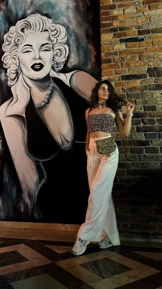 A young woman with long dark hair and light skin posing against a brick wall with a large black and white Marilyn Monroe portrait painting behind her. She is wearing a leopard print crop top, white pants, and sneakers, holding a small bag and touching her hair.