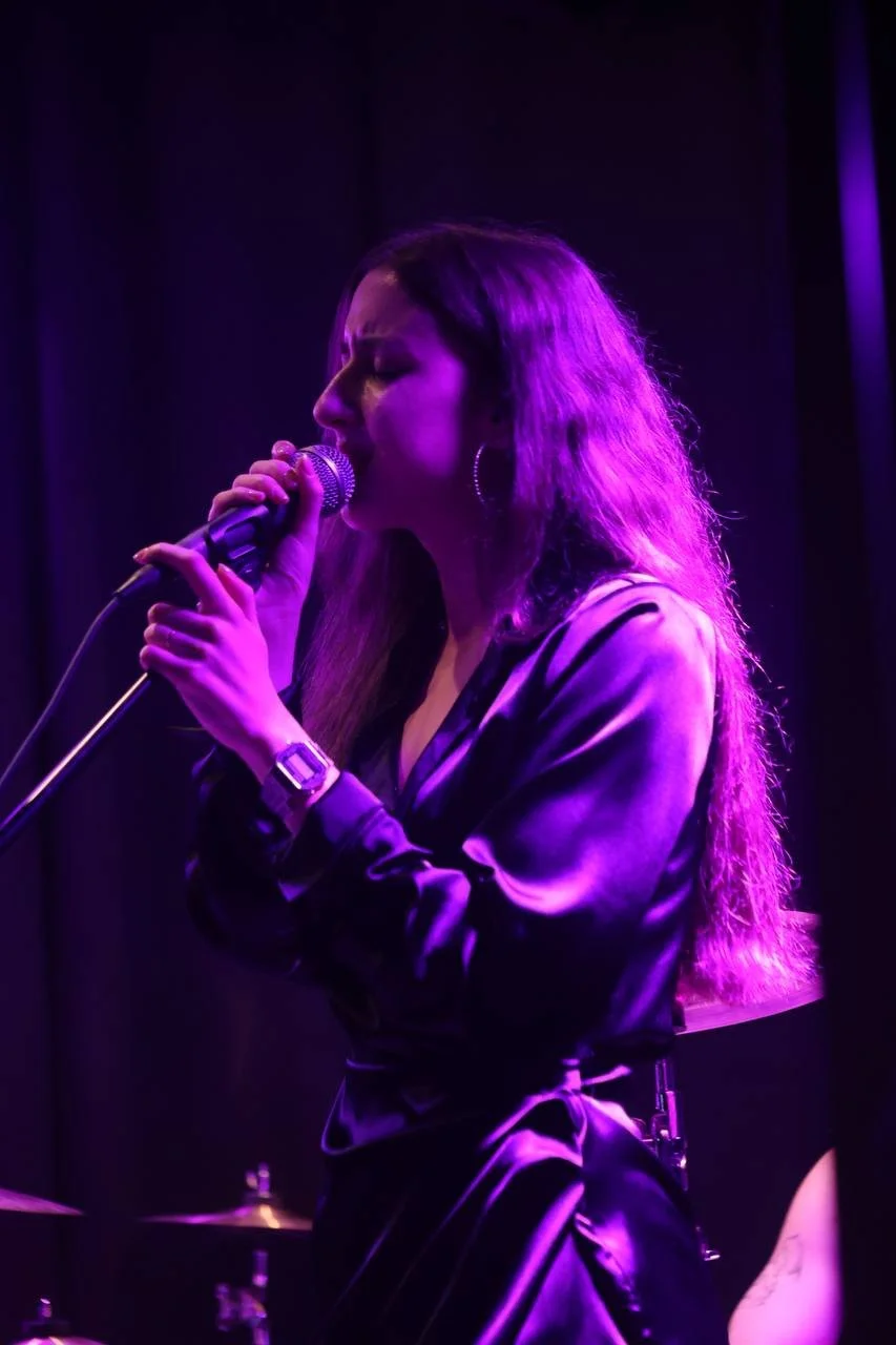 Female singer performing with a microphone on stage, illuminated by purple stage lighting.