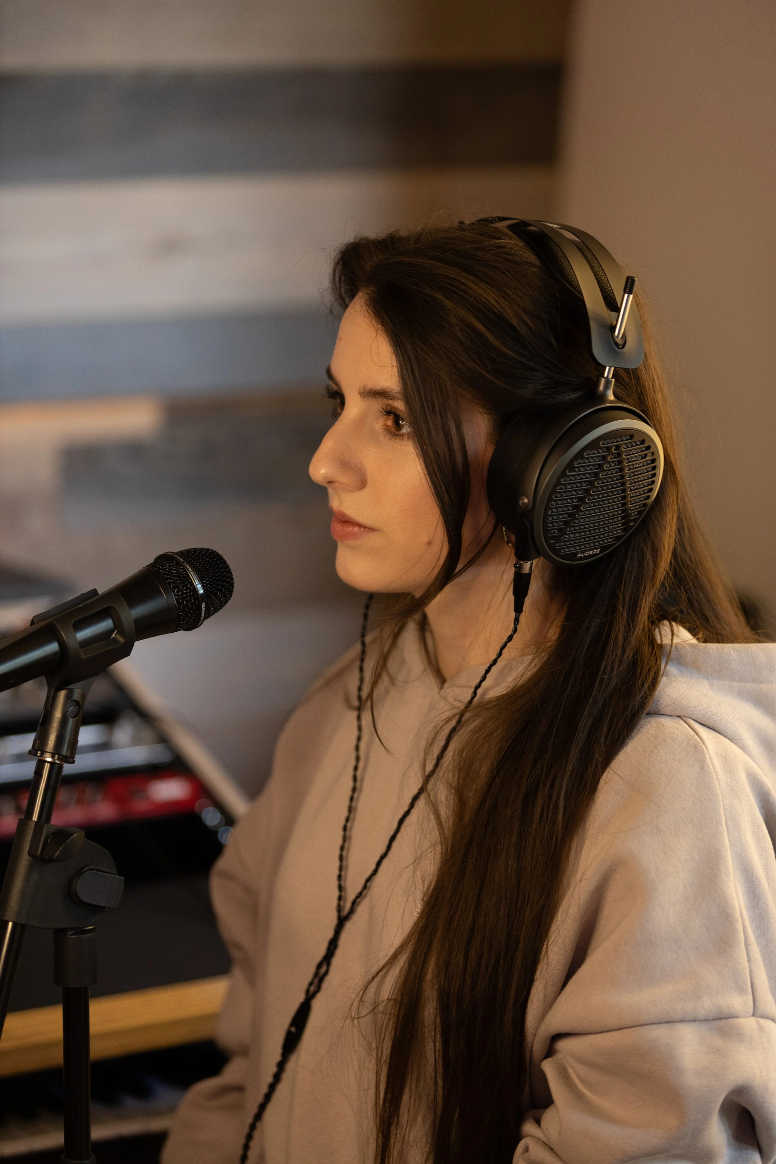 A woman wearing headphones and a beige hoodie singing into a microphone in a recording studio.