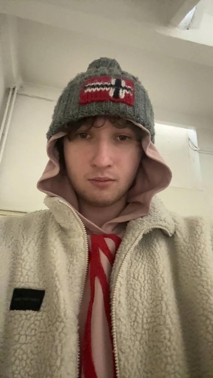 A young man wearing a gray beanie with a red and black design, a pink hoodie, and a beige fleece jacket, indoors.
