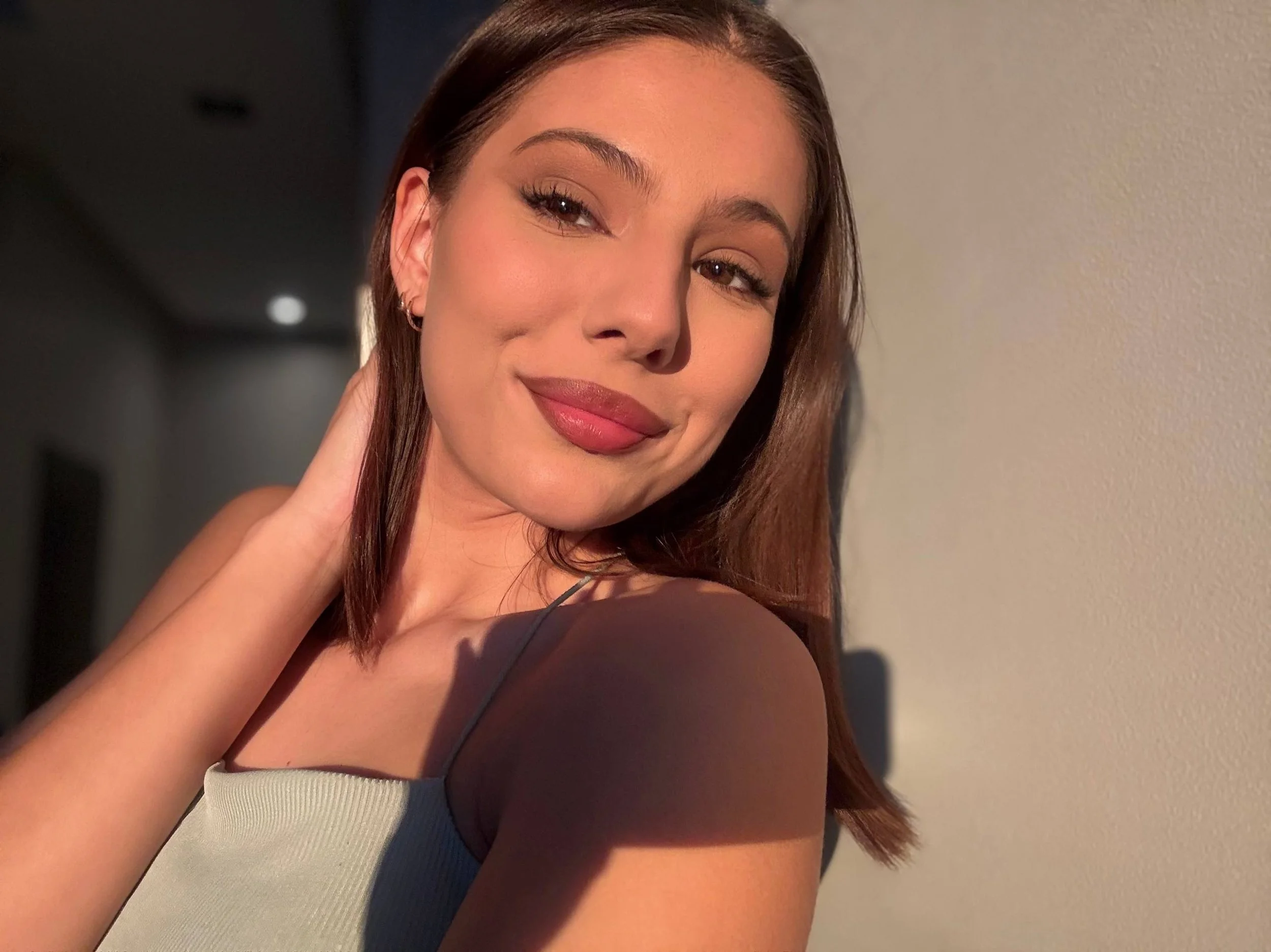 A young woman with brown hair, wearing a light-colored spaghetti strap top, is smiling softly while touching her neck, standing against a white wall with warm lighting.