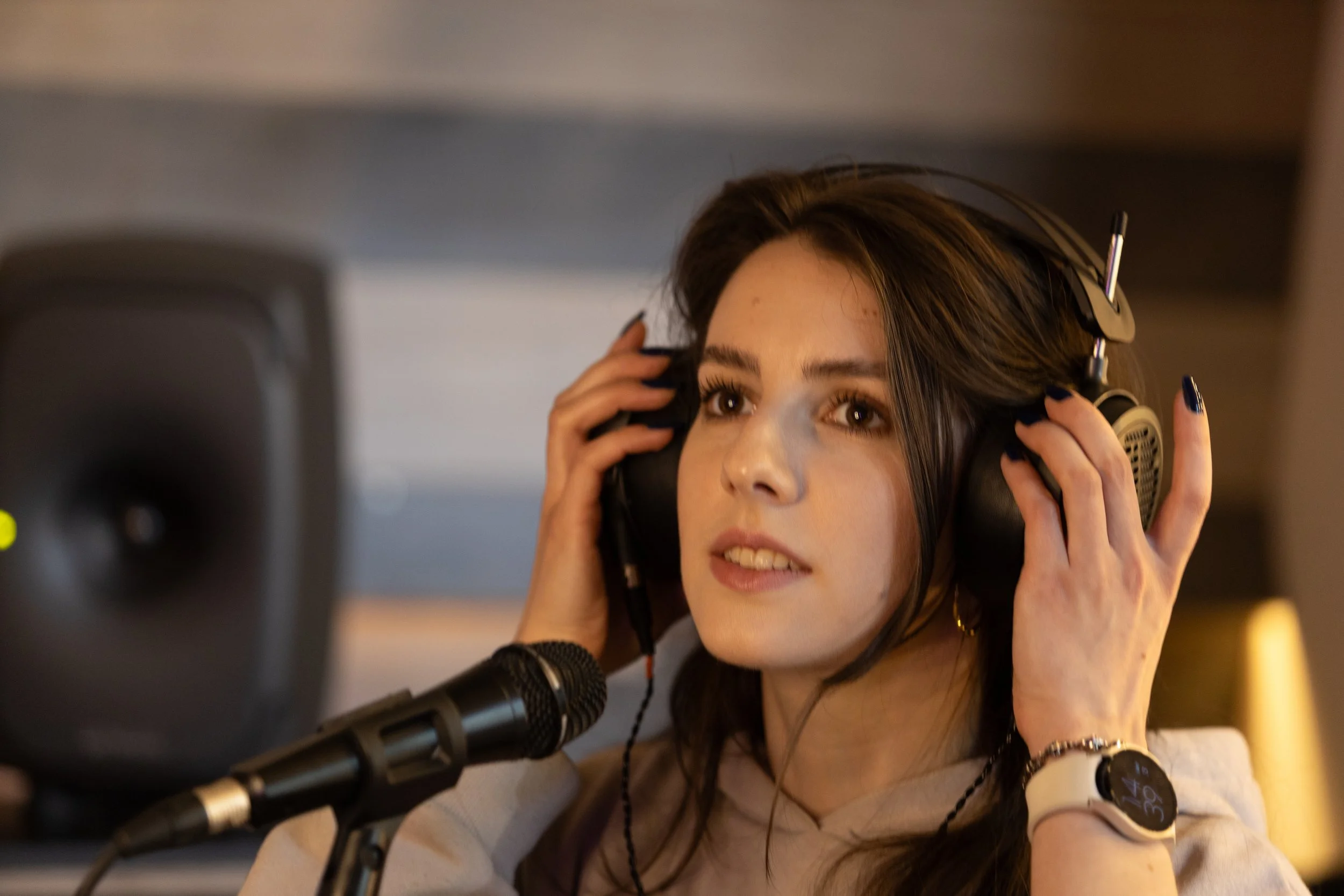 Young woman with dark hair wearing headphones, singing or speaking into a microphone in a recording studio.