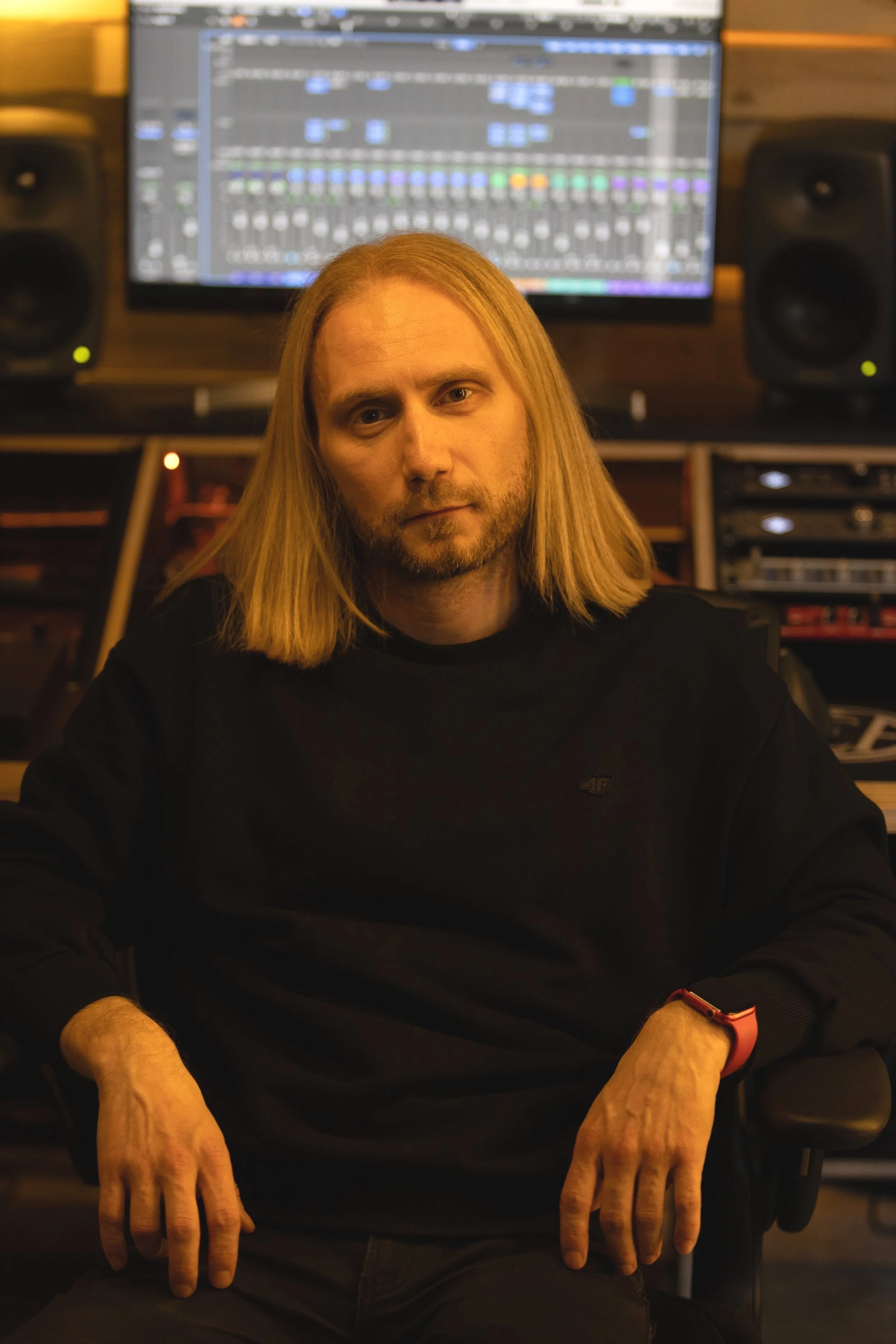 A man with long blond hair and a beard sitting in a recording studio with sound equipment and a computer screen displaying a music production software.