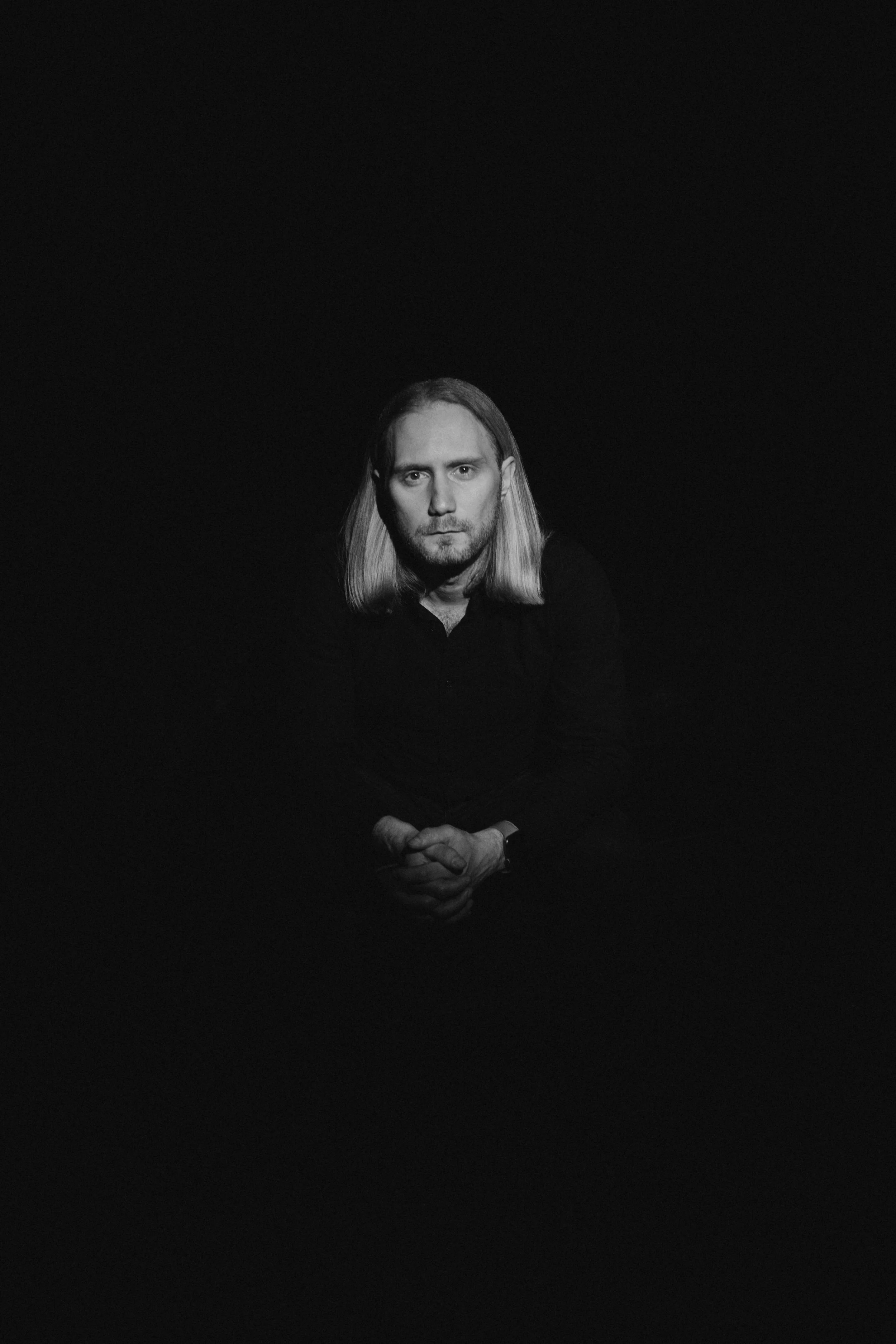 A black and white portrait of a man with long blonde hair, sitting with hands clasped, against a dark background.