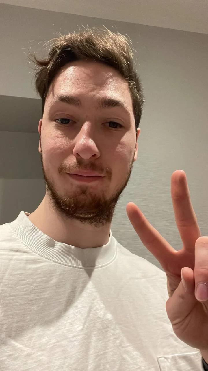 A young man taking a selfie indoors, making a peace sign with his right hand, with a slight smile on his face, wearing a white t-shirt.