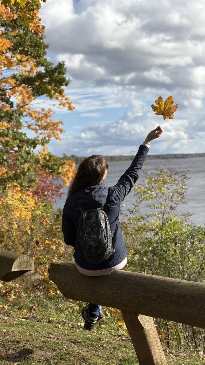 A woman sitting on a wooden fence near a lake or river, surrounded by autumn trees with colorful leaves, tossing a large yellow leaf into the air on a cloudy day.
