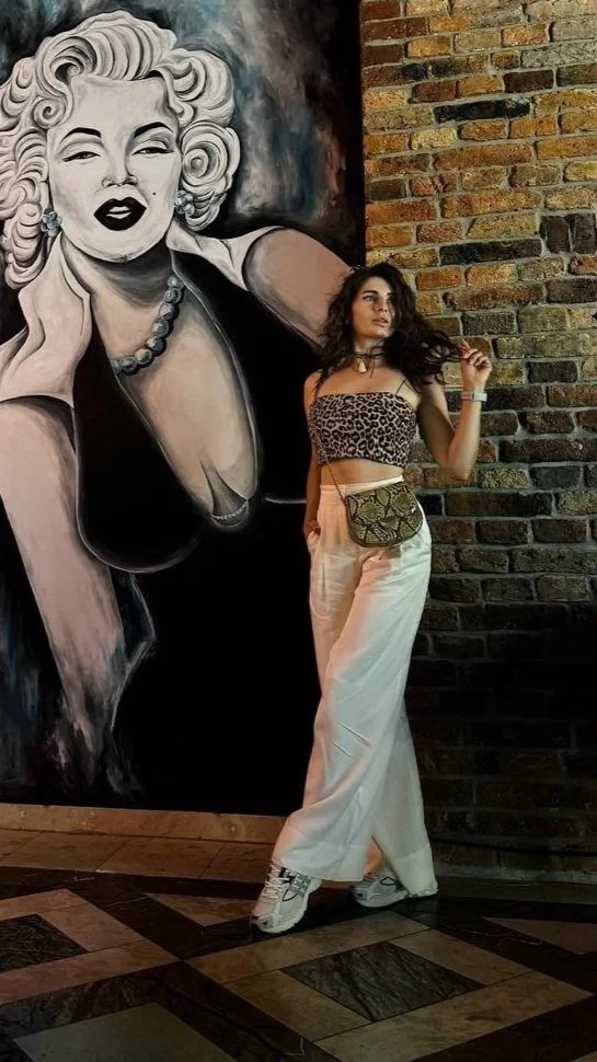 A woman with long dark hair, wearing a leopard print crop top, wide-leg white pants, and sneakers, stands next to a large black-and-white mural of Marilyn Monroe on a brick wall.