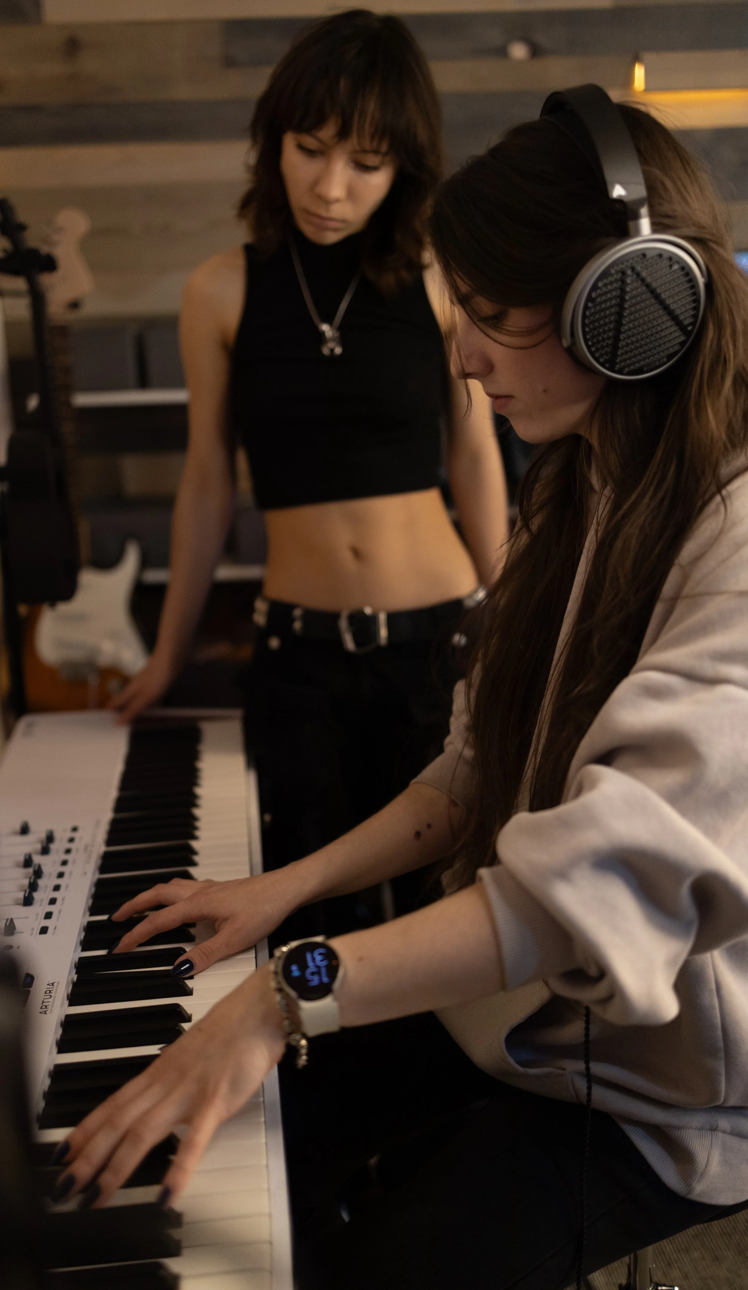 Two young women in a music studio, one playing a MIDI keyboard and the other standing nearby. The woman playing the keyboard wears headphones and a beige hoodie, while the other woman wears a black crop top.