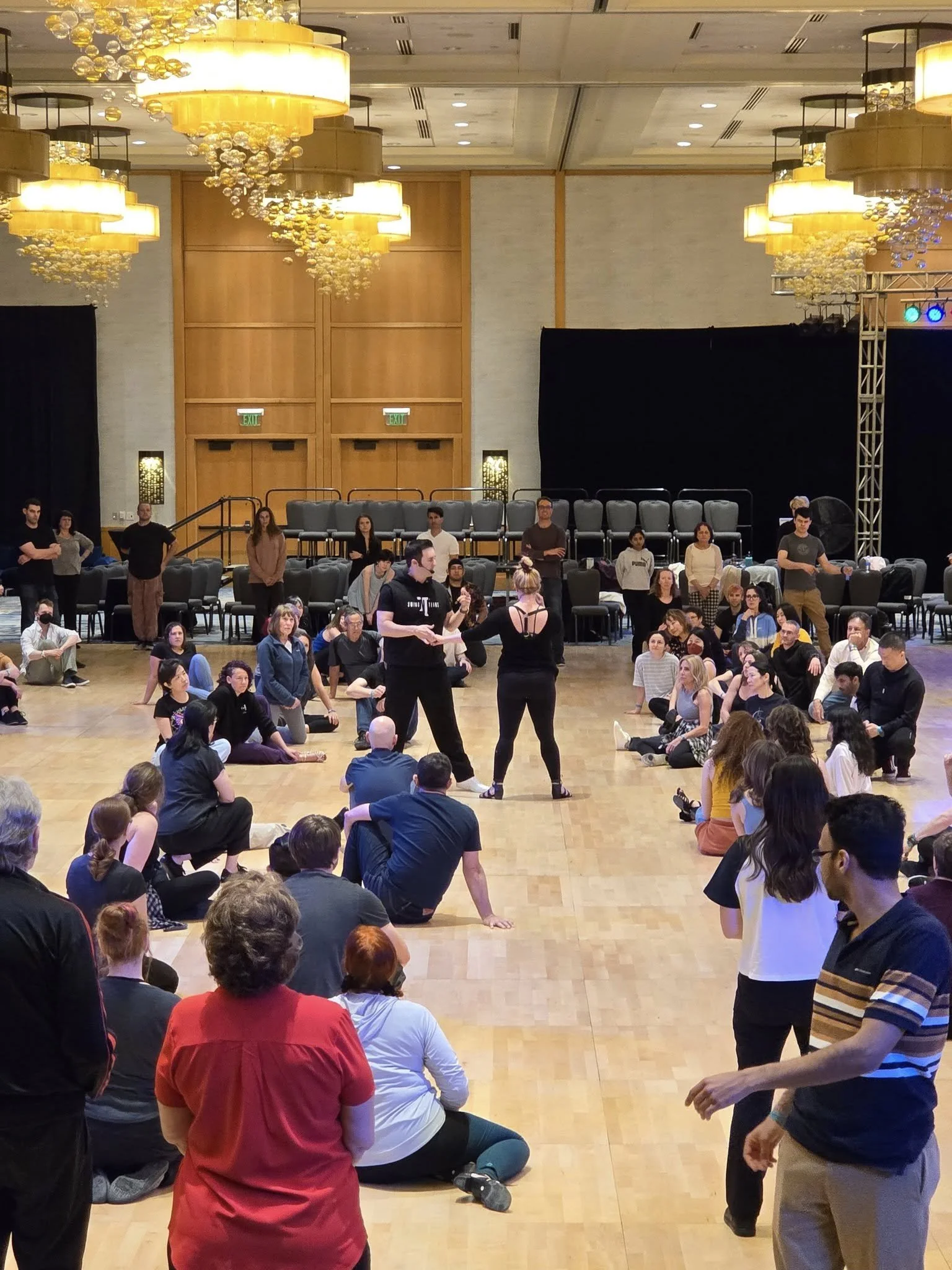 People seated in a large ballroom watching a dance rehearsal or performance on a wooden floor with two dancers in the center, surrounded by audience members sitting on the floor and chairs, with large chandeliers hanging from the ceiling.
