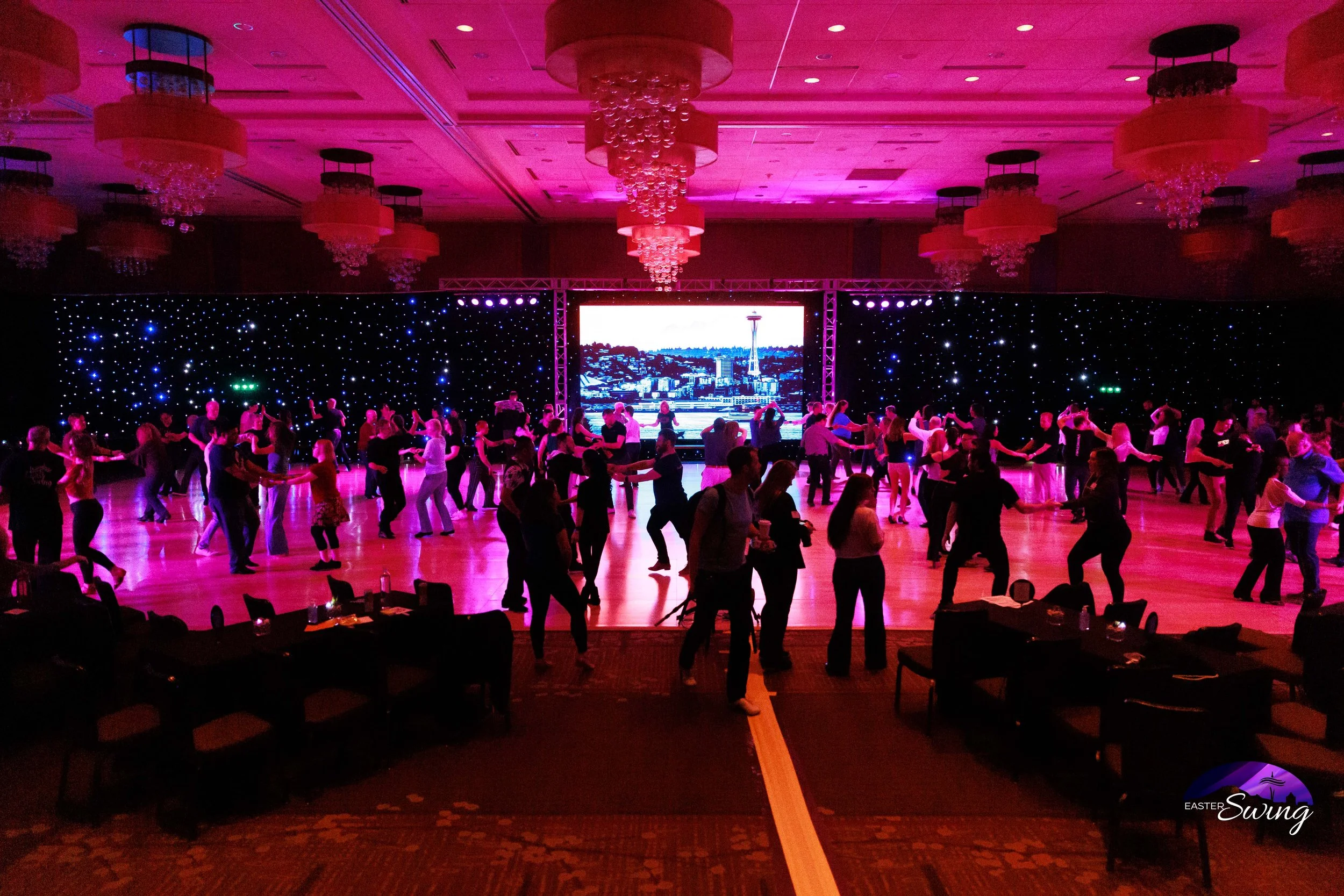 People dancing in a large ballroom with pink lighting, a starry backdrop, and a large screen displaying the Seattle Space Needle.