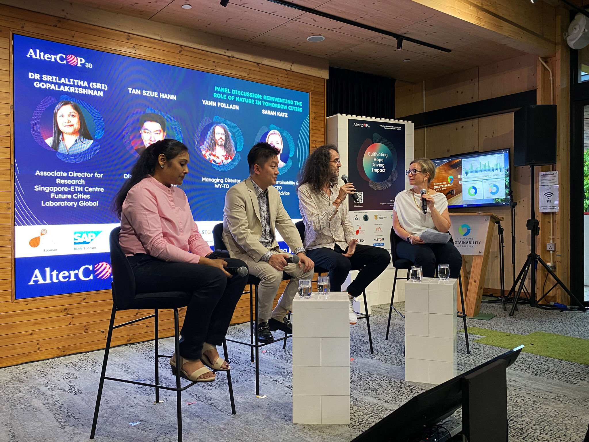 A panel at a conference, four panelists are sitting, with Sarah Katz to the right with a microphone in her hand.