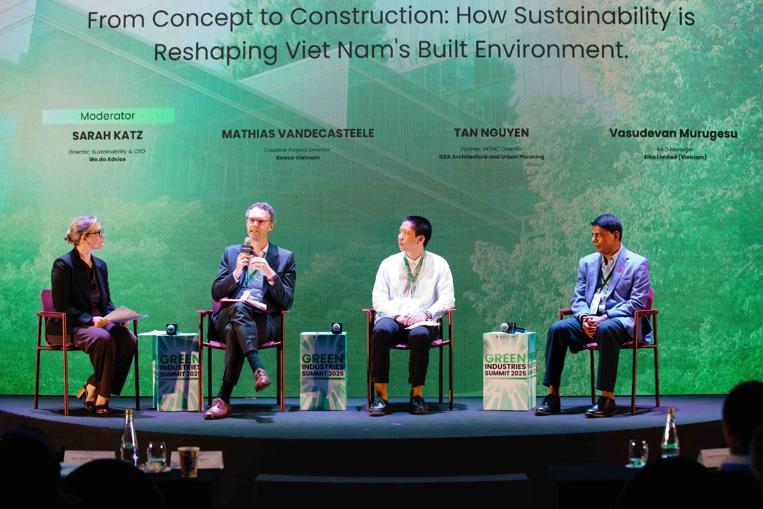 A panel at a conference, Sarah Katz is sitting to the left, her name and the word moderator is displayed above her on the wall. Two other panelists are on the right.