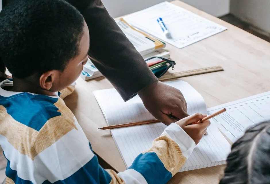 A teacher leaning over a wooden desk to point at an open notebook while assisting a young student with their schoolwork