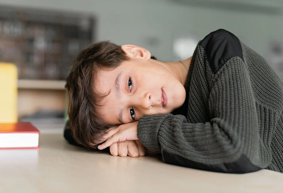 A young boy rests his head on a table, looking thoughtfully into the distance.