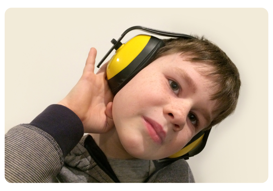 7-year old boy wearing a gray and black stripped hoodie and bright yellow noise-cancelling headphones turning his head and holding his right ear.