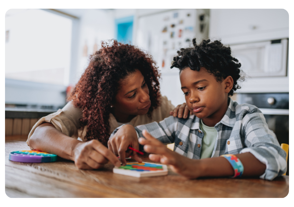 Eight-year old African American boy sitting at a table doing a colorful puzzle with the assistance of his mother, who has her hand on his shoulder and is talking to him.