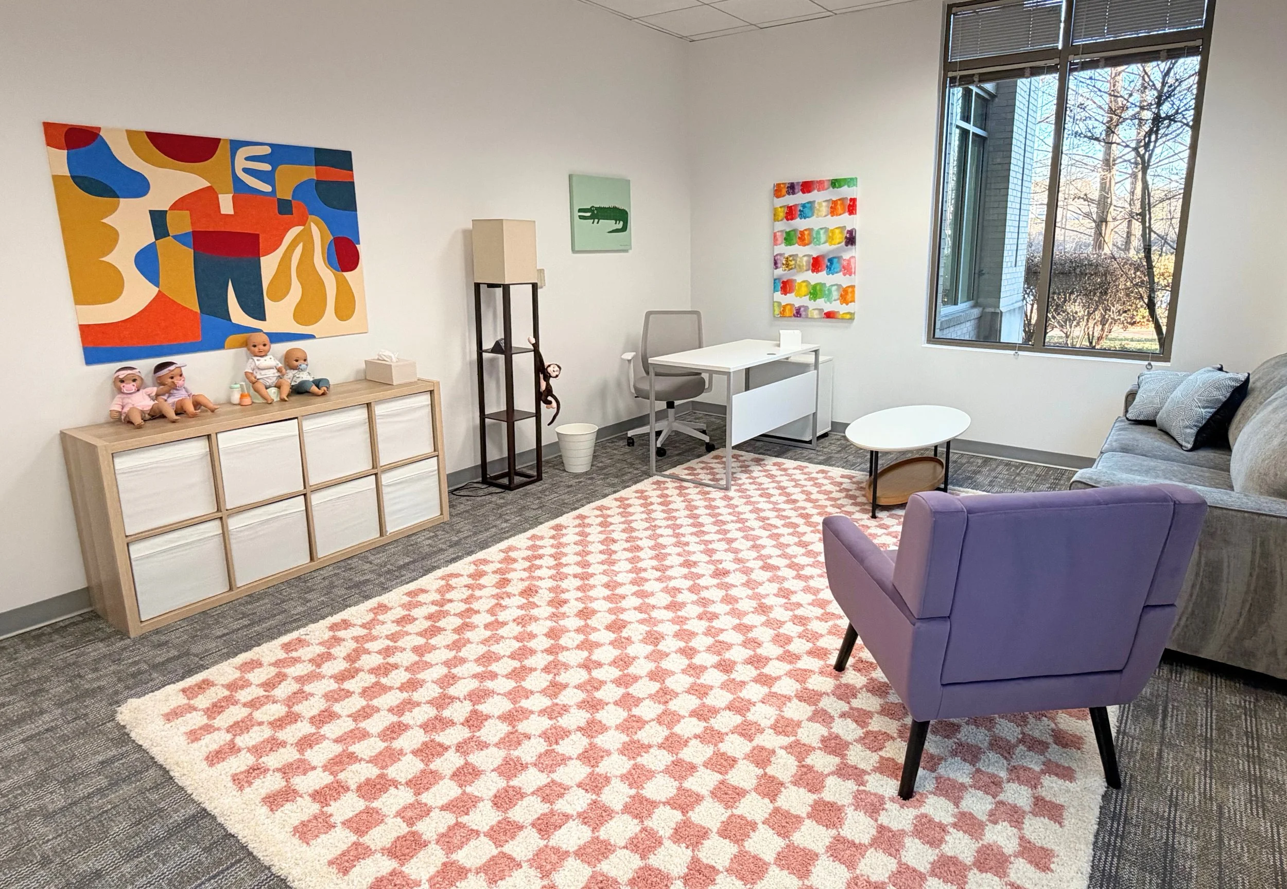 A cheerful white meeting room with a pink and cream checkerboard area rug, purple armchair, gray couch, colorful abstract art, and window looking out onto trees.