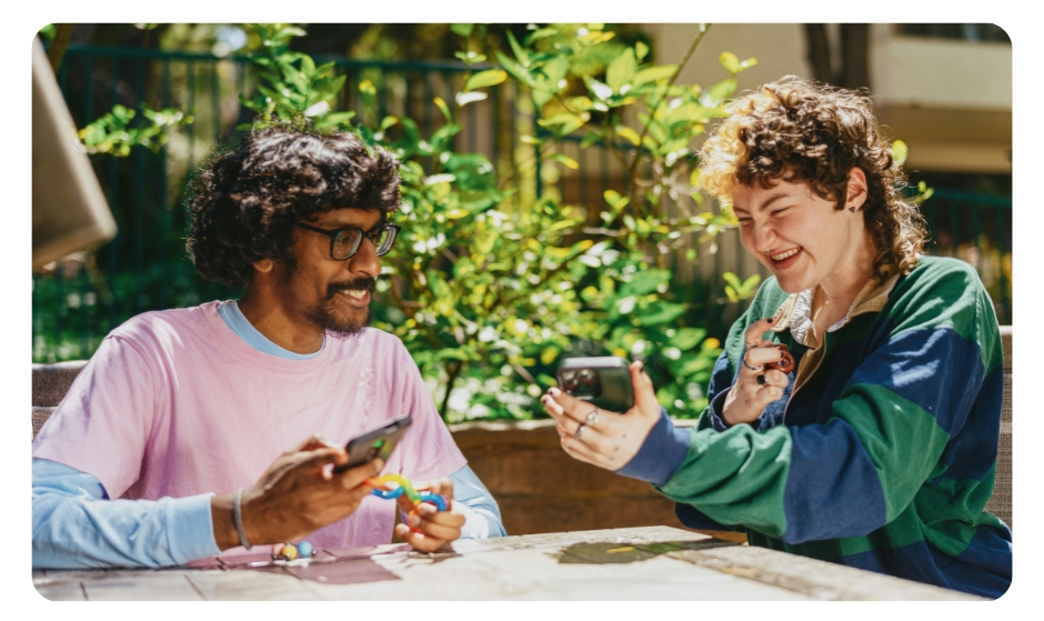 A teenage girl wearing a blue and green striped rugby shirt laughing and showing her phone to a teenage boy wearing glasses. Both are holding colorful puzzles.