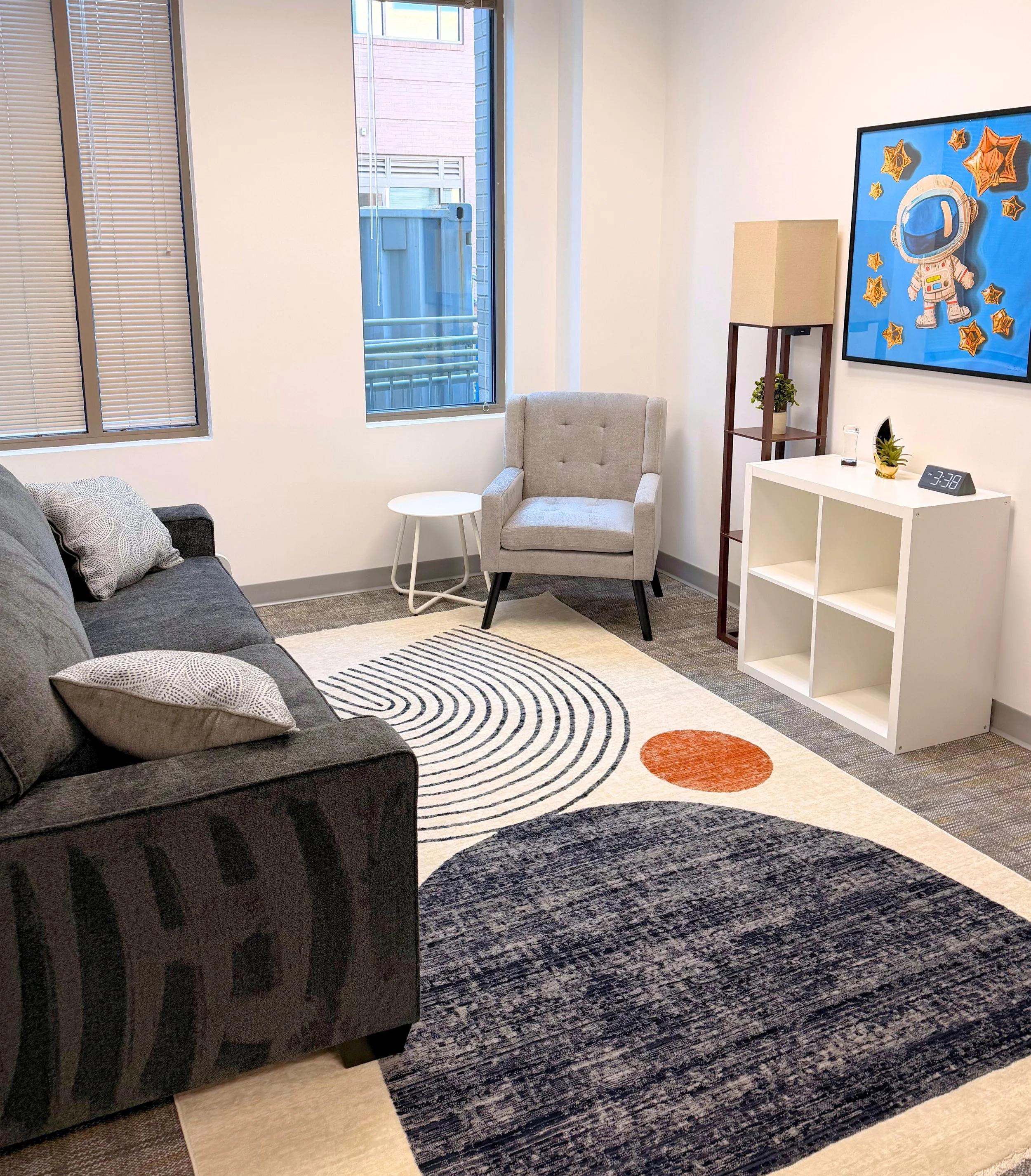 Cheerful white meeting room with area rug depicting abstract landscape and sun. Gray couch, gray armchair, white shelf cubbies, and wooden floor lamp with shelves. 
