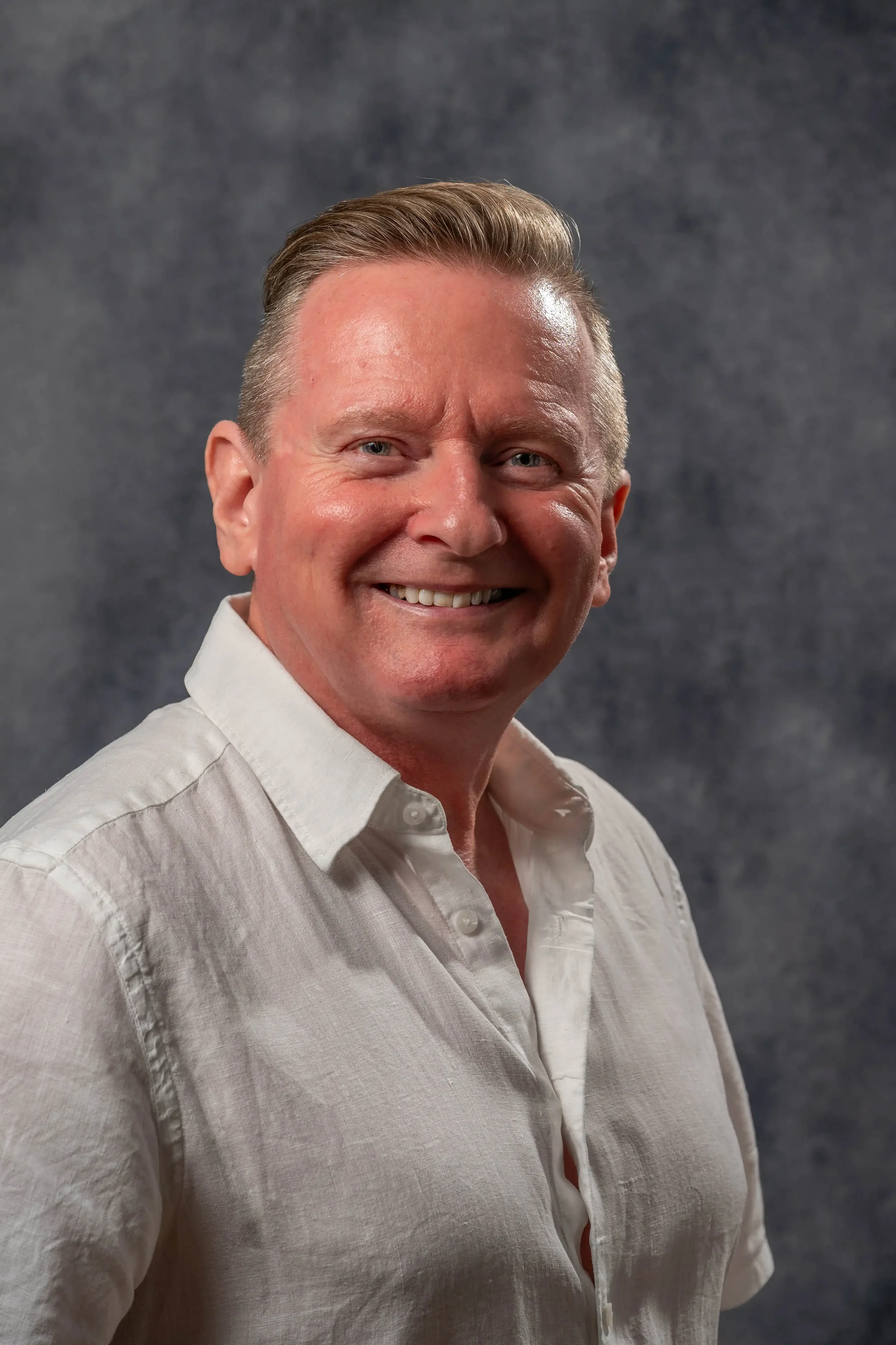 Headshot photograph of a man with fair hair, wearing a casual white shirt, against a dark grey background