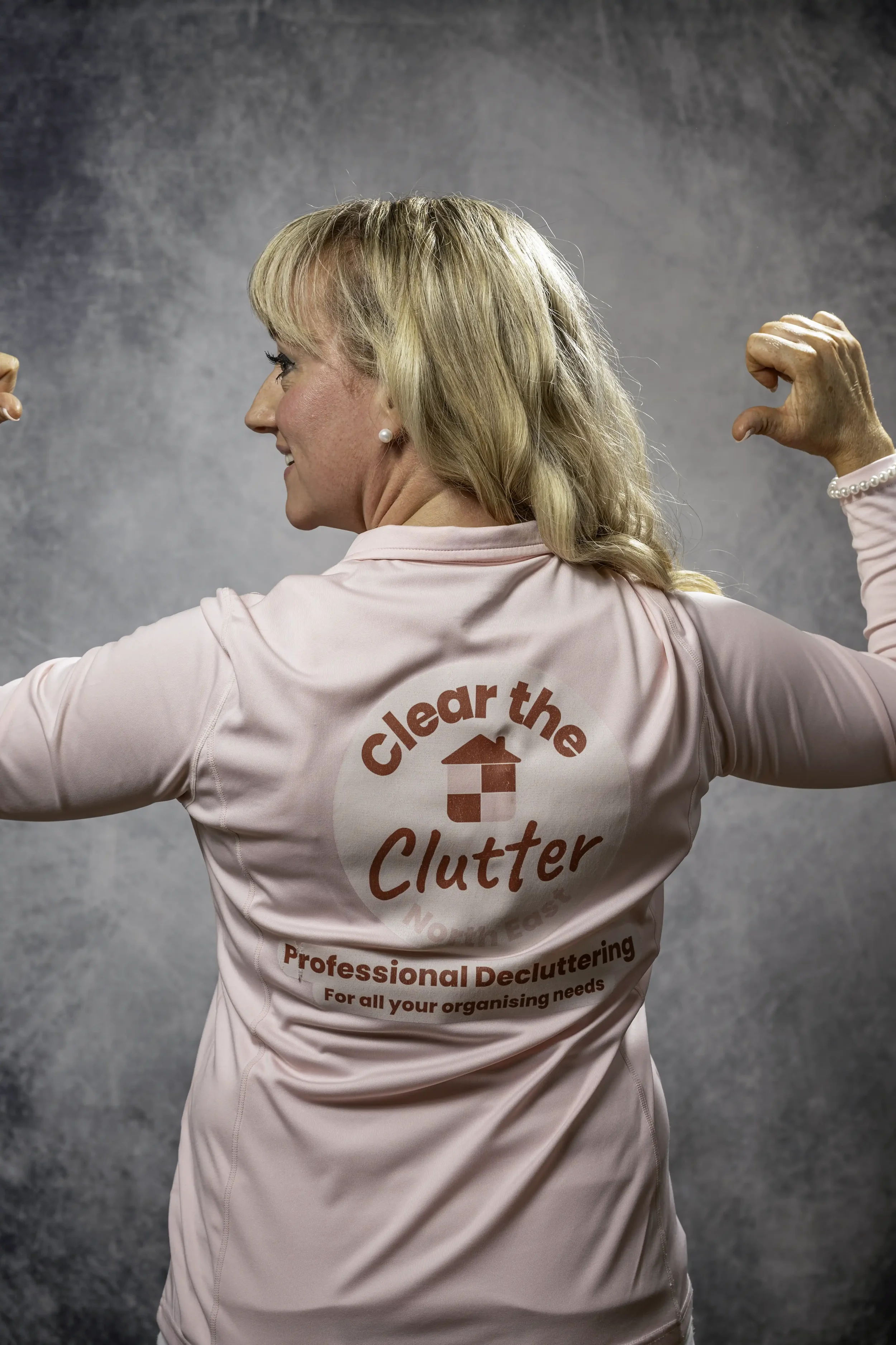Photograph of a woman with blonde hair in a long-sleeved pink top with a company logo, standing with her back to the camera, with her arms raised and pointing at her company logo, standing in front of a mid-grey backdrop