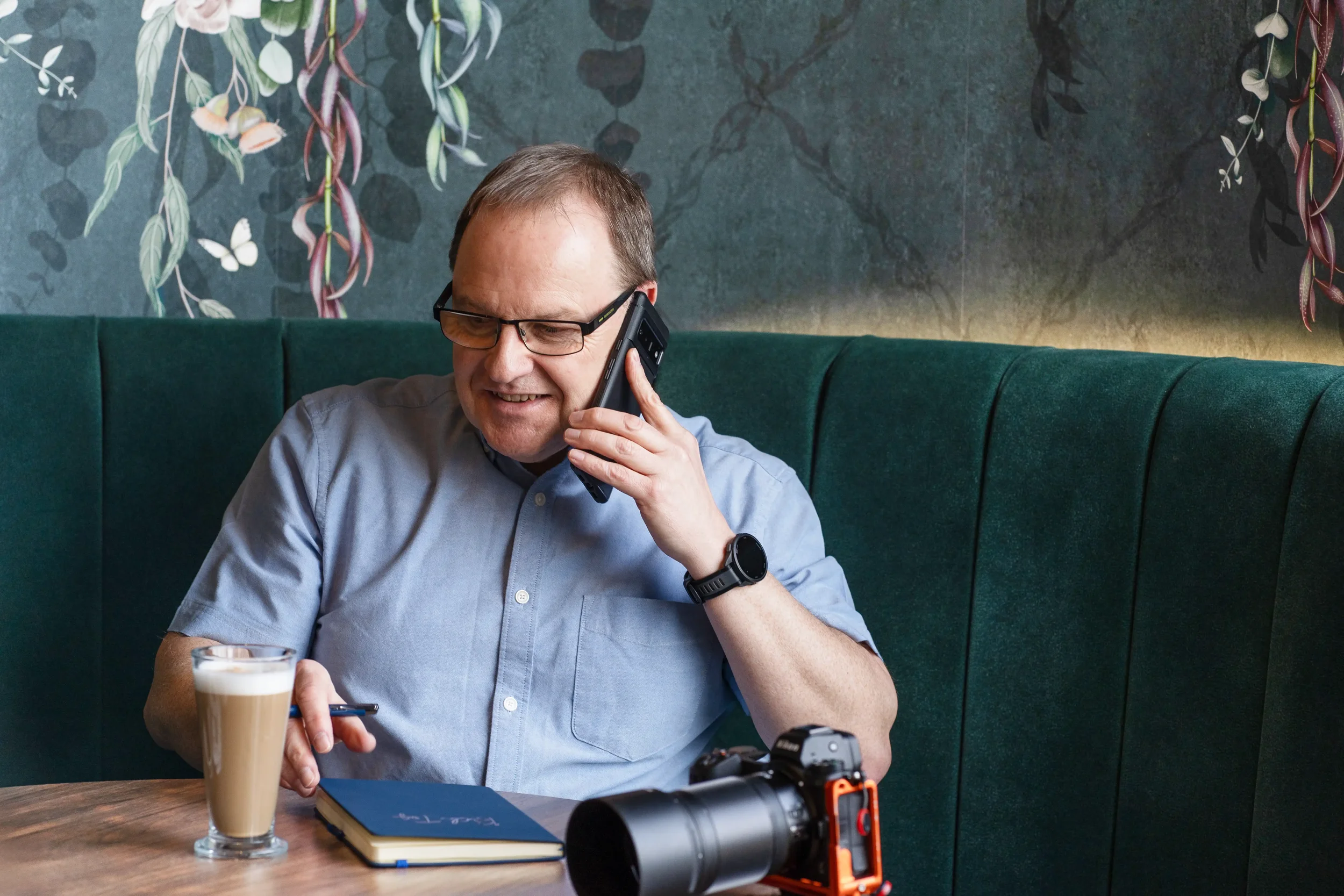 Colour portrait of a man with short brown hair, wearing a light blue shirt, sitting in a hotel taking a phone call, with a coffee and a camera beside him, sitting on a green seat with green patterned wallpaper in the background.