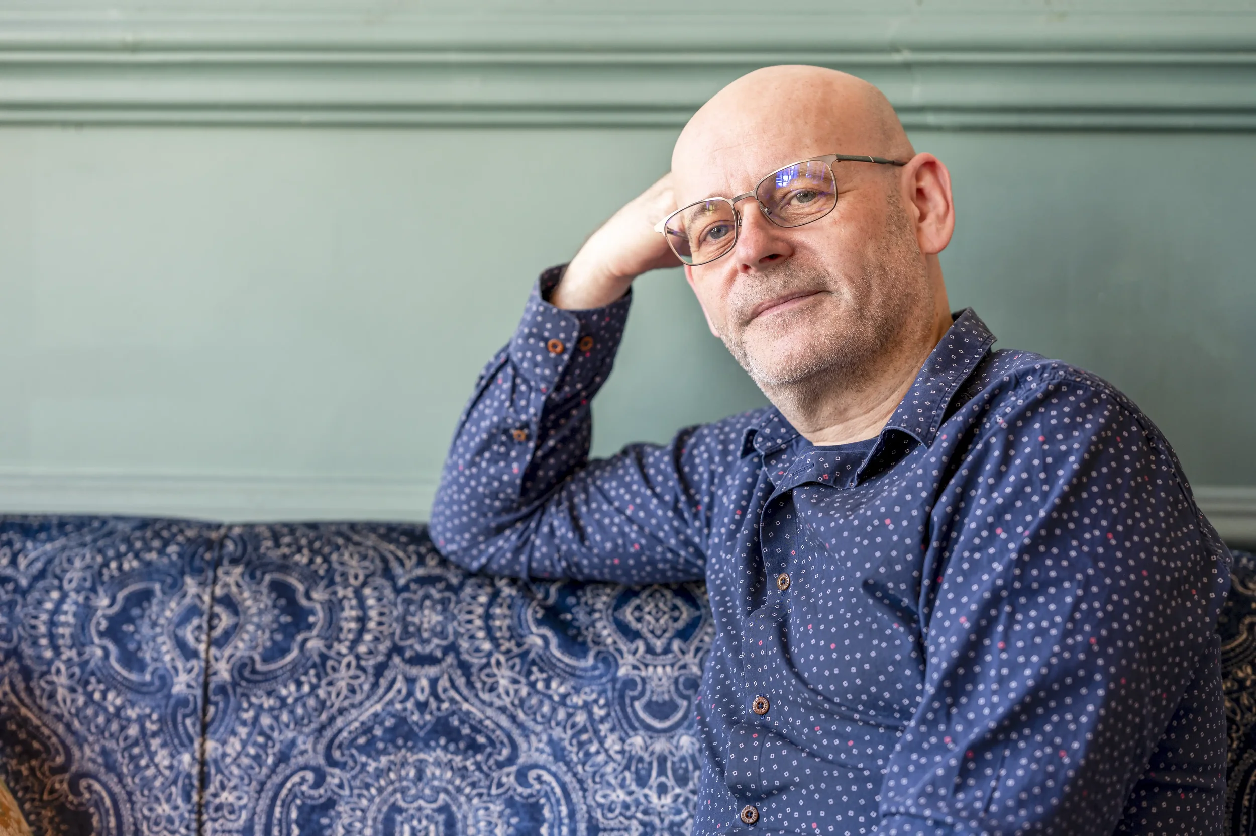 Personal brand image of a man wearing a blue shirt sitting on a blue patterned sofa, against a green panelled wall.