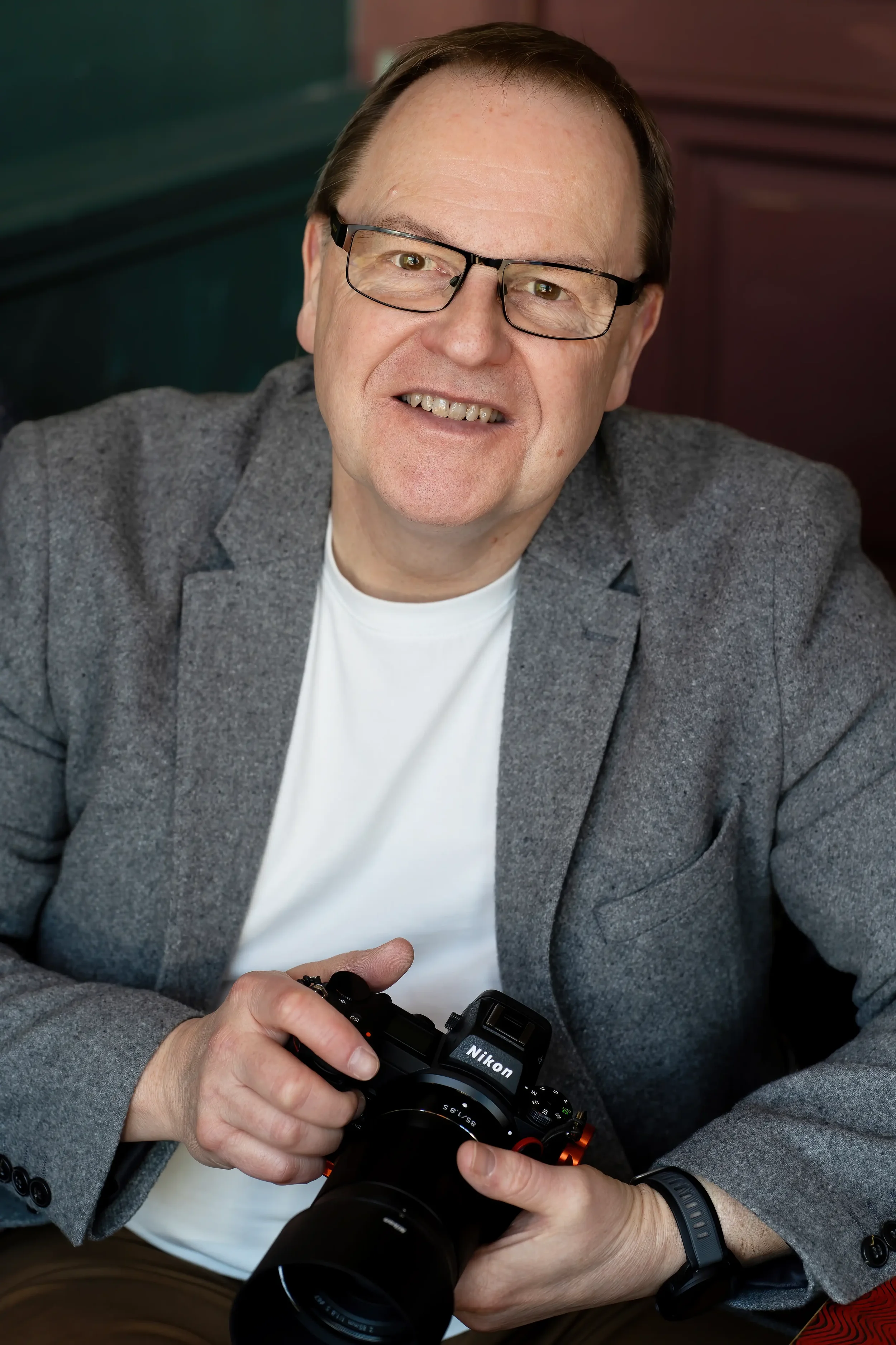 Colour portrait of a smiling man with glasses and short brown hair, wearing a white T-shirt, grey jacket and brown chinos, holding a camera, relaxing in a cafe, with a green and a pink wall in the background.