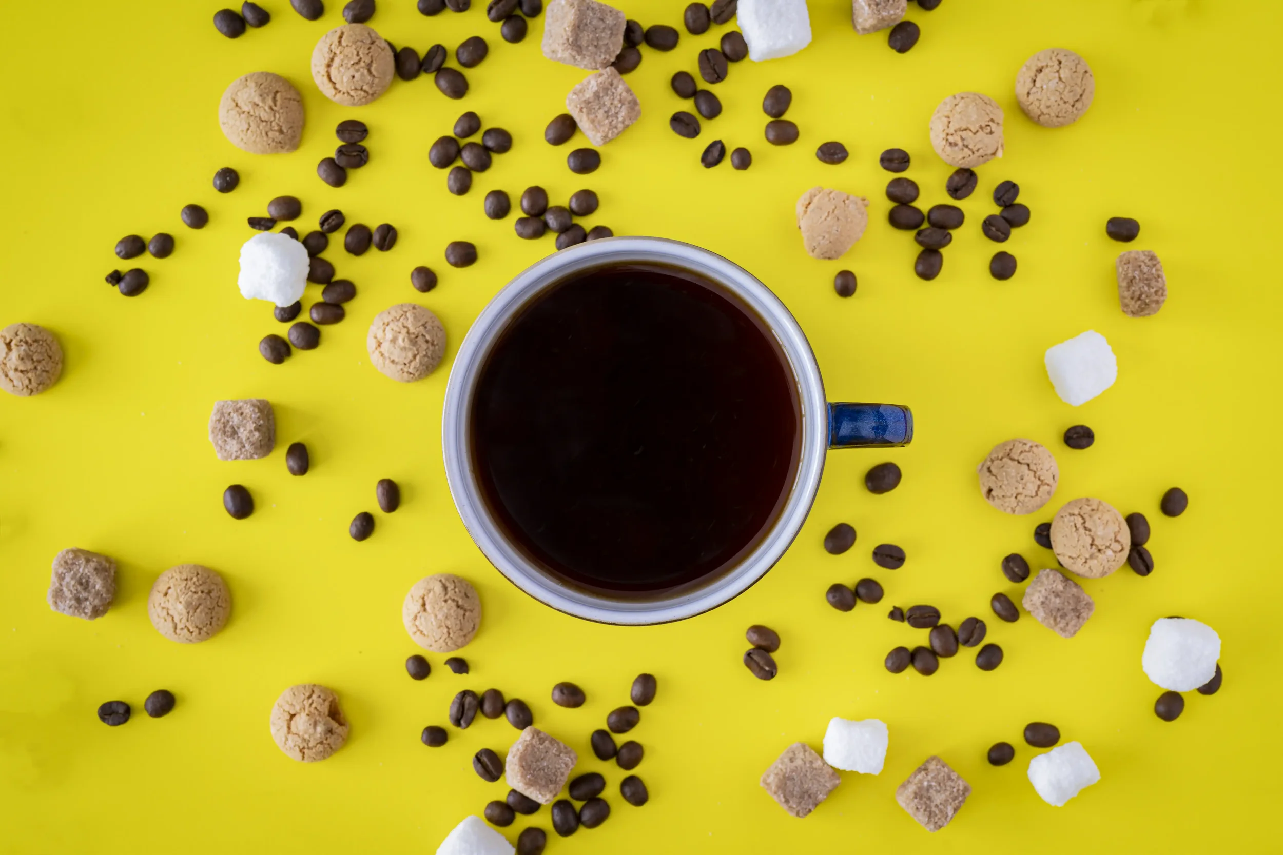 Flat lay colour photograph looking straight down on a cup of coffee on a bright yellow background, surrounded by coffee beans, amaretti biscuits and brown and white sugar cubes