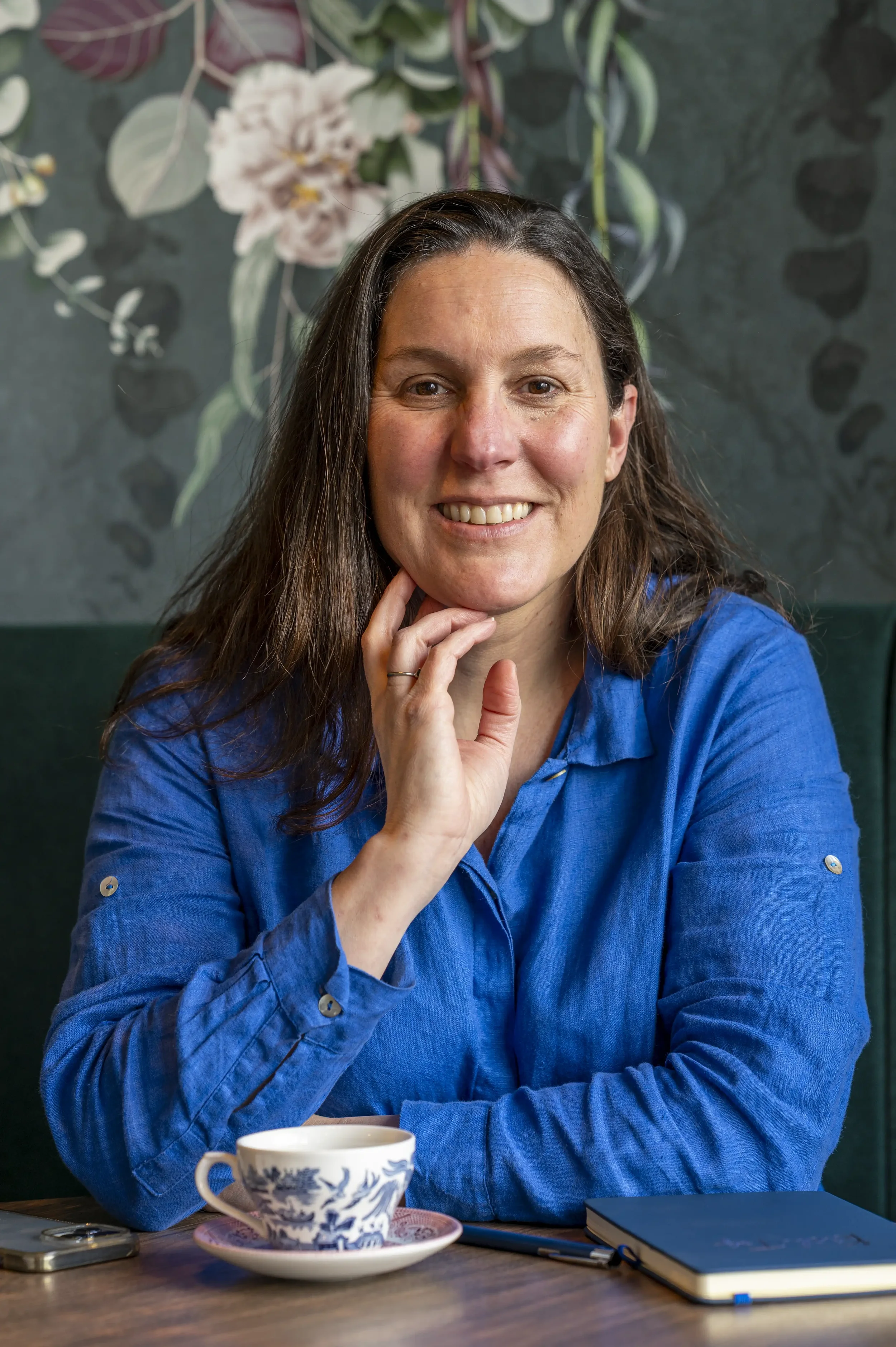 Headshot photograph of a smiling woman with long black hair wearing a blue shirt, with a cup of coffee in a cafe with green floral wallpaper in the background