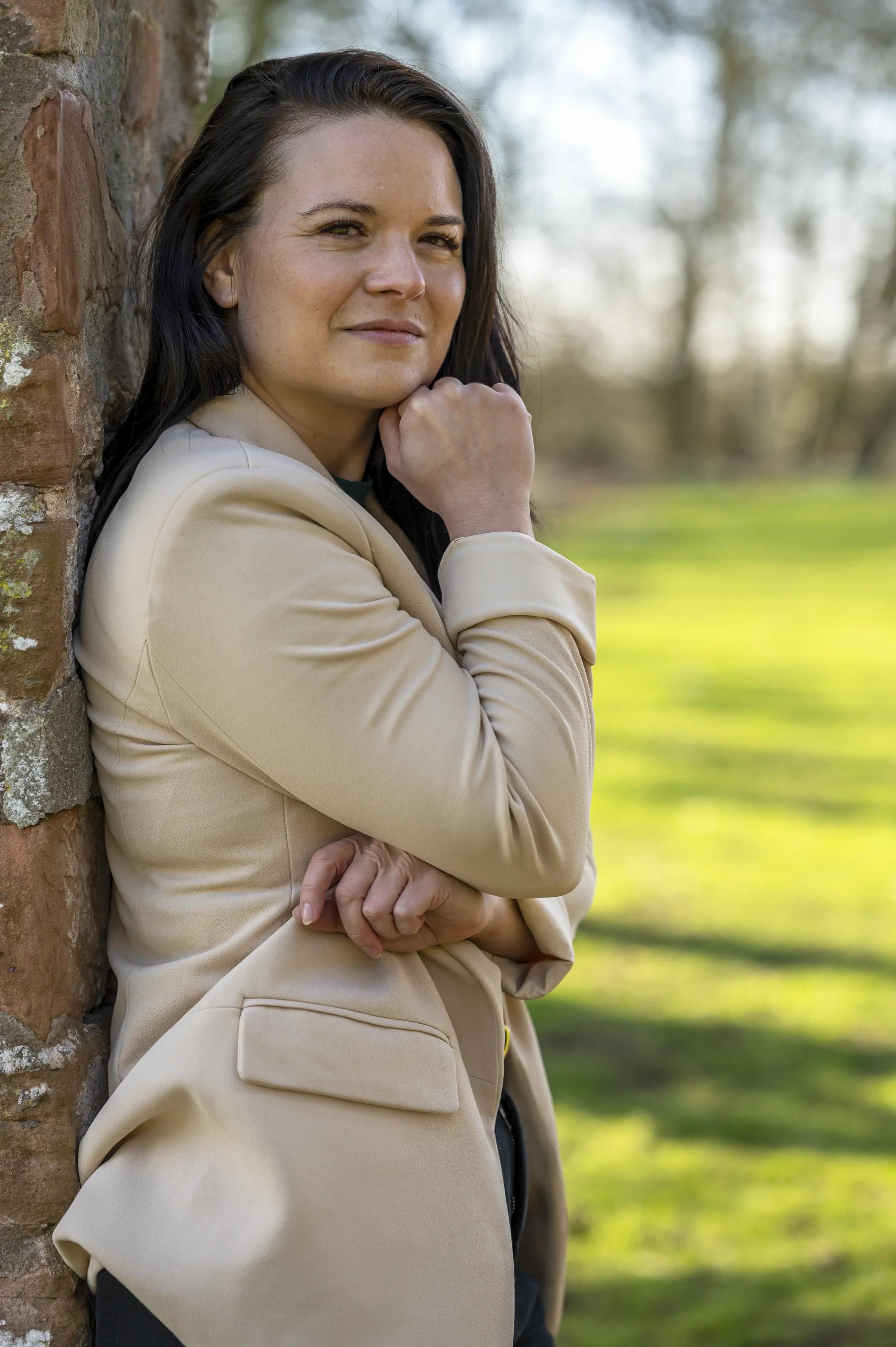 Headshot photograph of a woman with long black hair wearing a green jumper and cream jacket, leaning against a brick wall, with trees and a field in the background