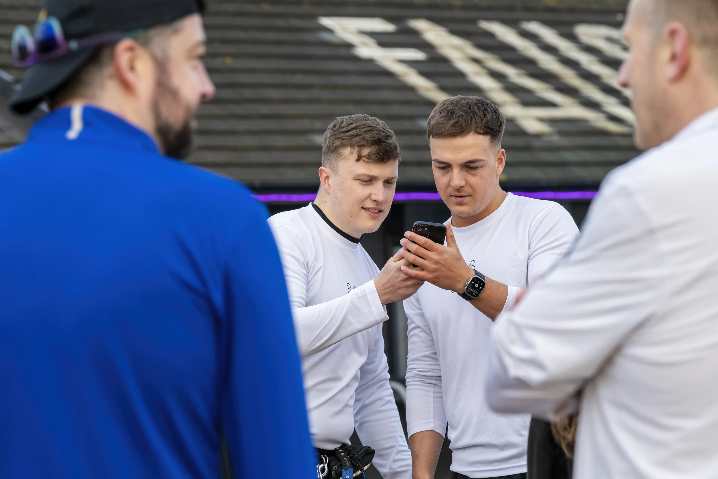 Photograph of two male runners wearing white long-sleeved running tops, looking at a mobile phone, taken through a gap between two other male runners in the foregrouund