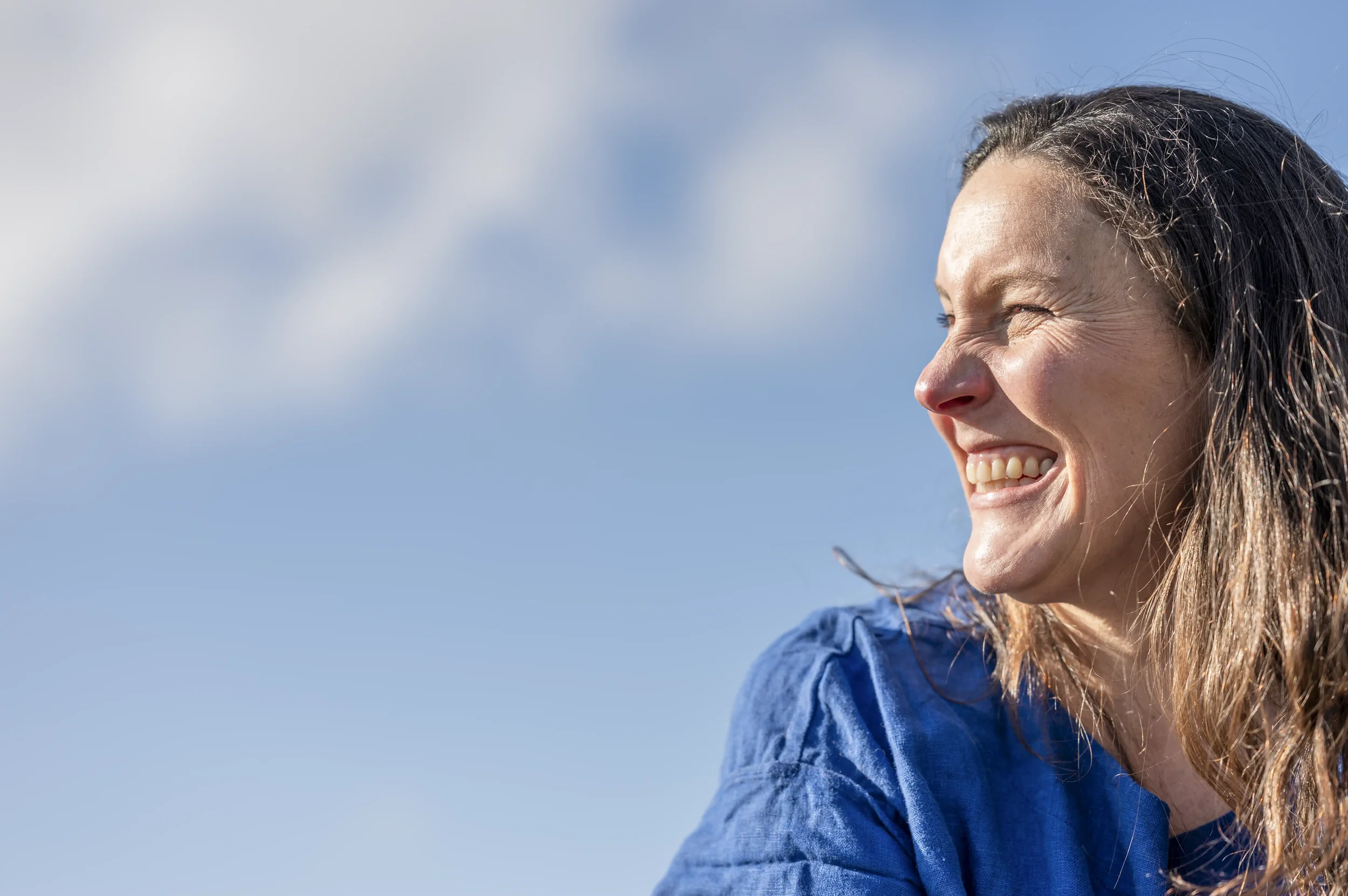 Headshot photograph of a smiling woman with long black hair wearing a blue shirt, against a blue sky with fluffy white clouds
