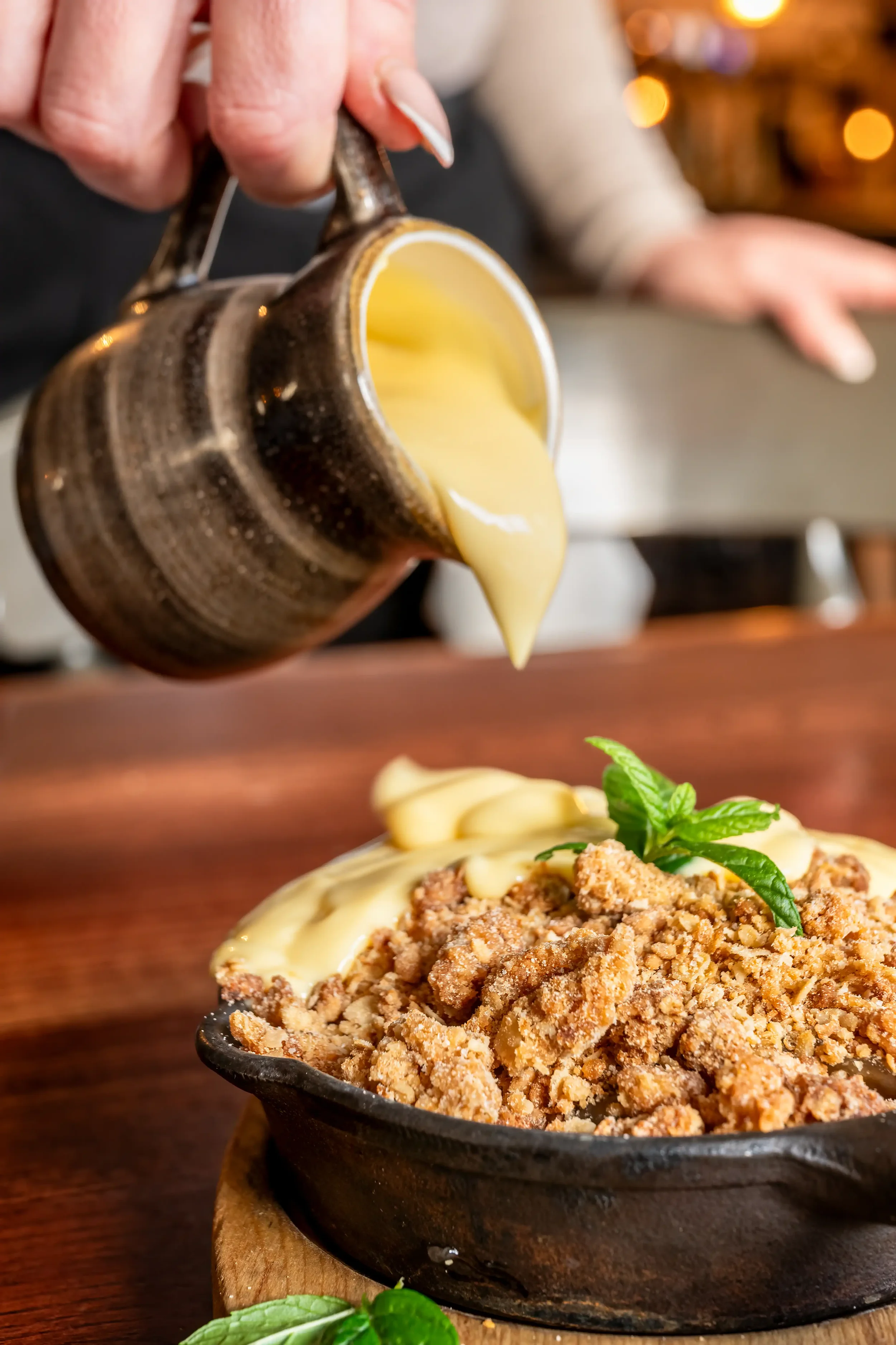 Colour photograph of a portion of apple crumble being served in a country pub, with fresh custard being poured on it from an earthenware jug