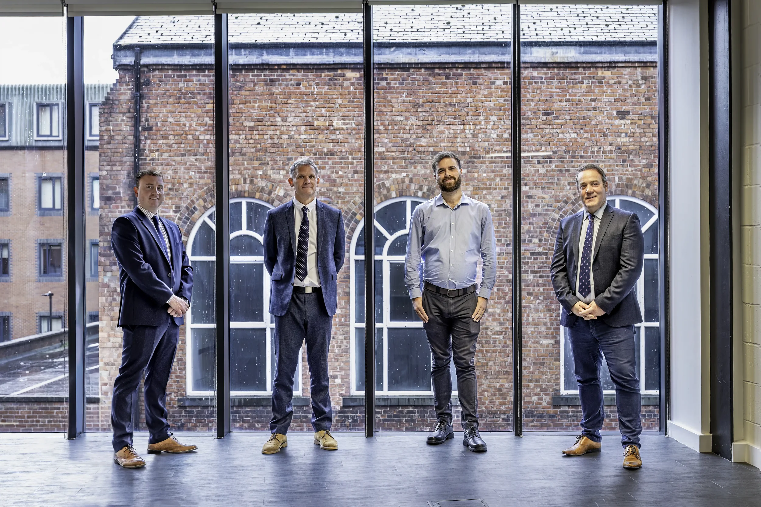 Colour photograph of four men in suits in an office standing in front of a lagre follow to ceiling window, with a red brick building in the background