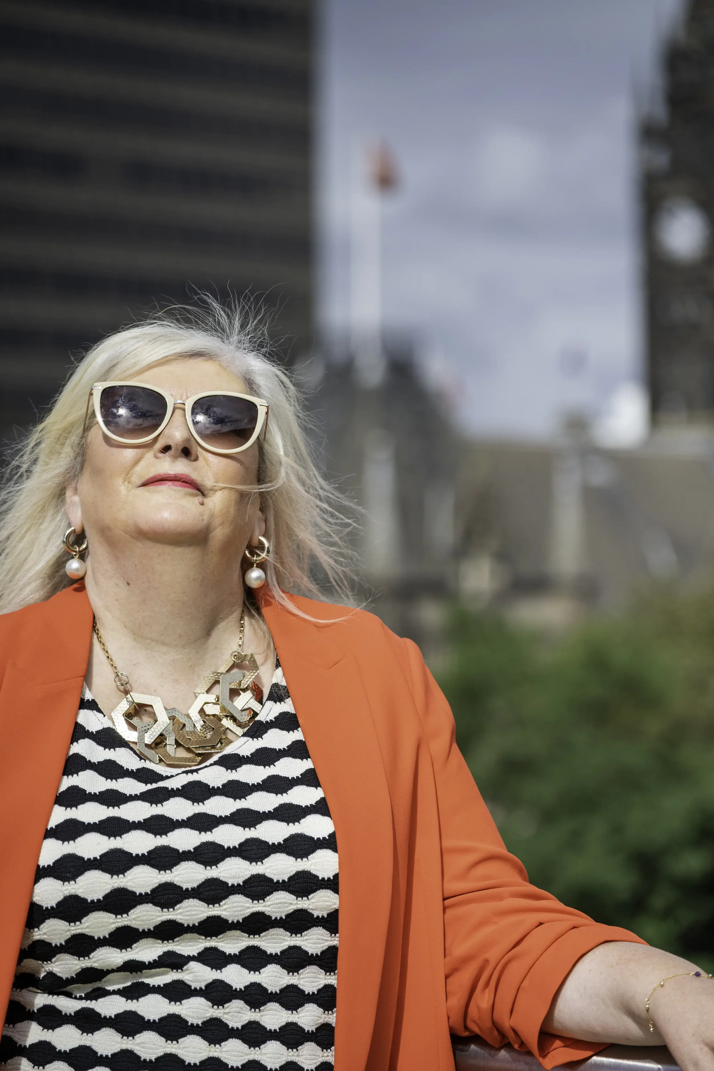 Photograph of a woman with blonde hair, wearing a black and white top, red jacket and sunglasses, staring up at the sun, with a city skyline in the background
