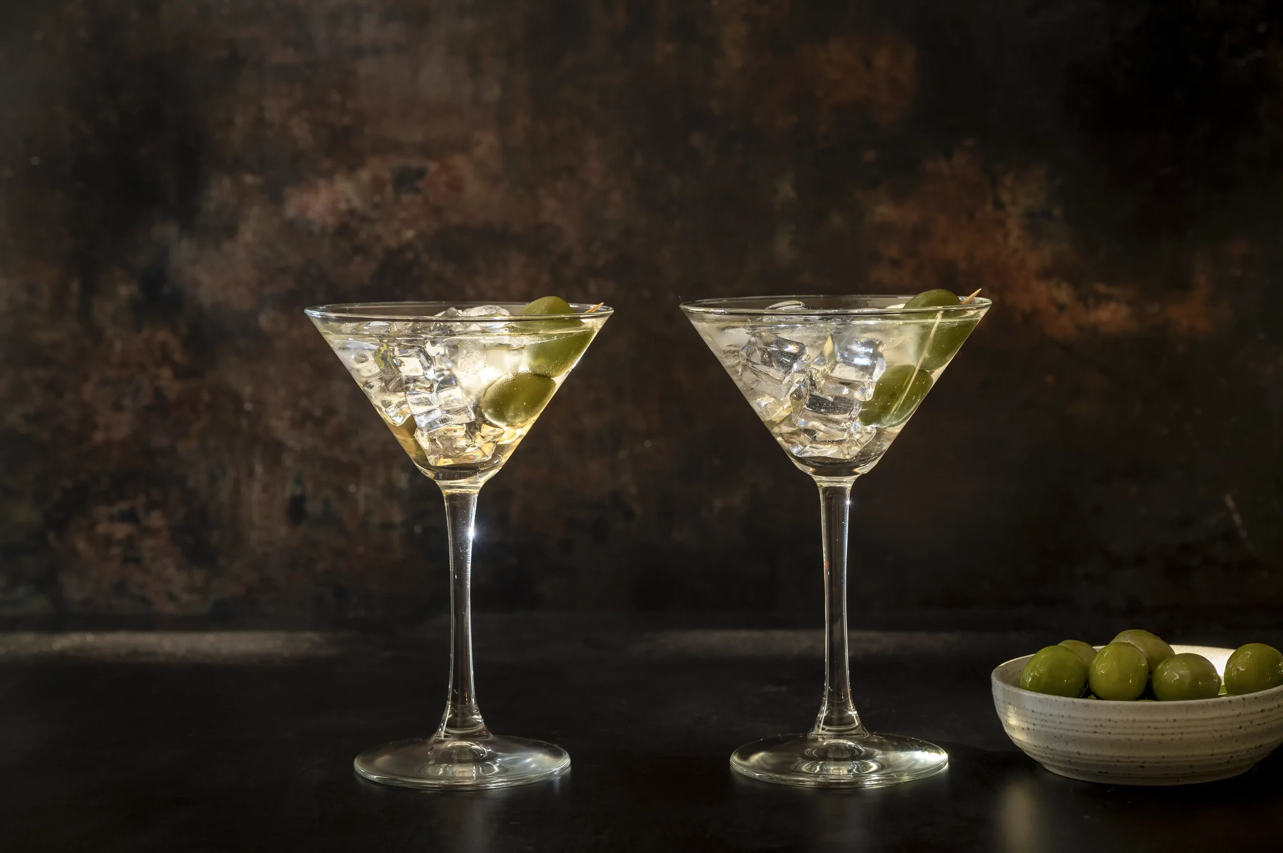 Image of 2 Martinis in conical glasses, with olives, set against a dark background.