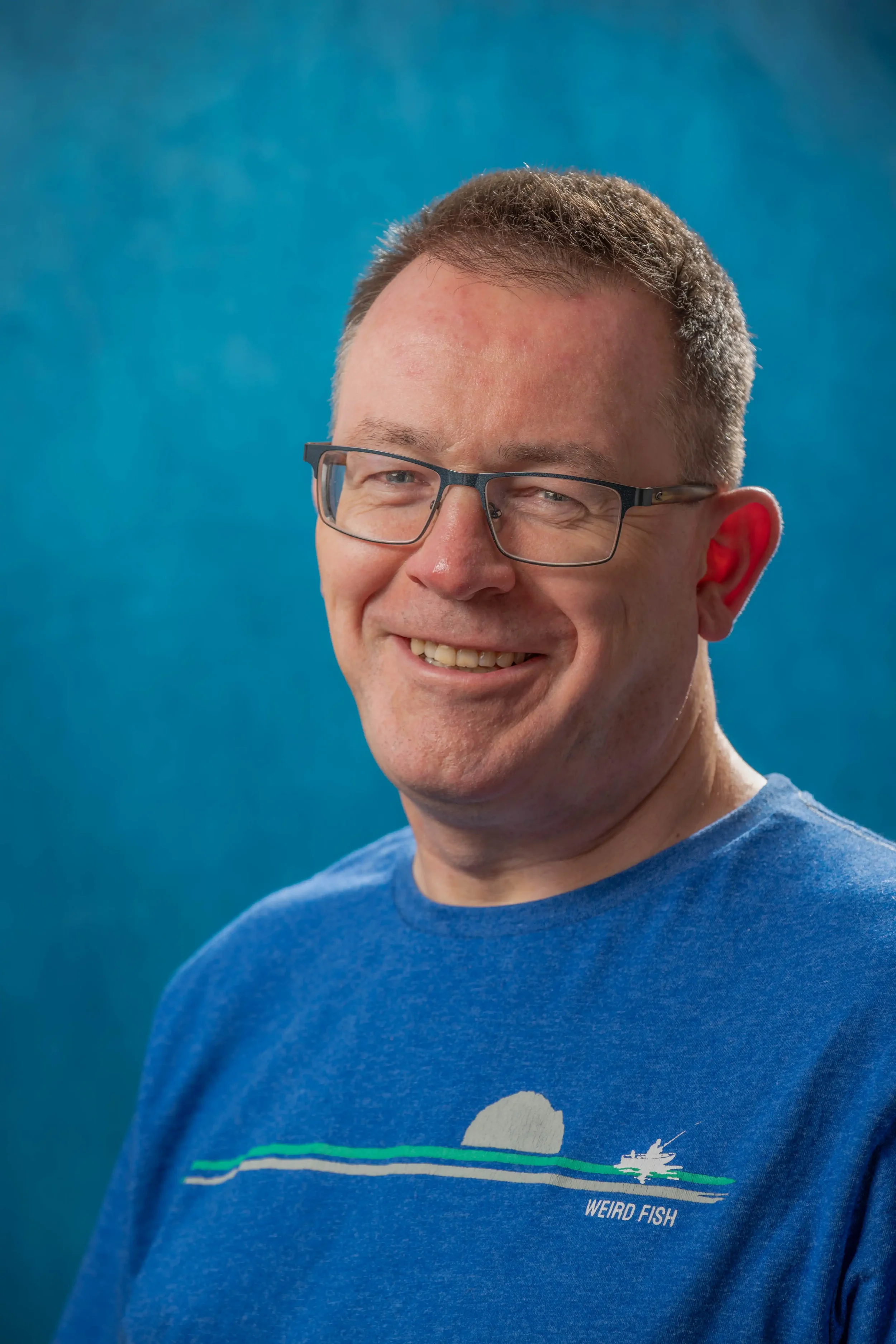 Headshot photograph of a man with glasses and short dark hair waering a blue T-shirt, against a blue background