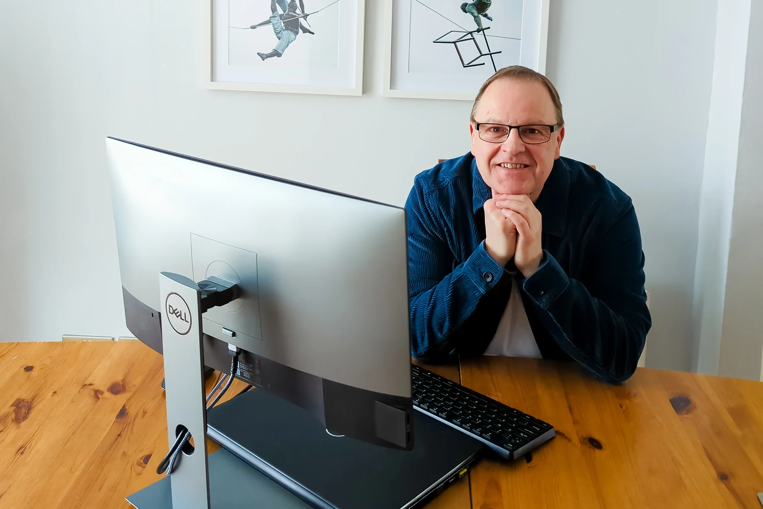 Colour portrait of a smiling man with glasses and short brown hair, wearing a white T-shirt and blue jacket, sitting at a table in front of a computer