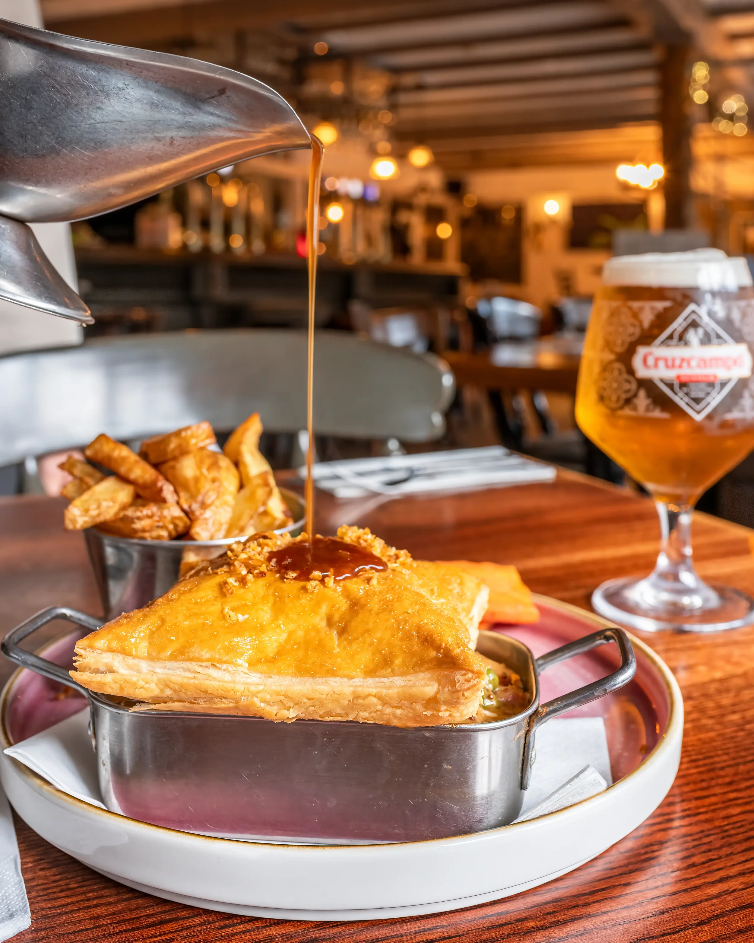Colour photograph of a pie being served in a traditional country pub, with gravy being poured onto it from a stainless steel gravy boat
